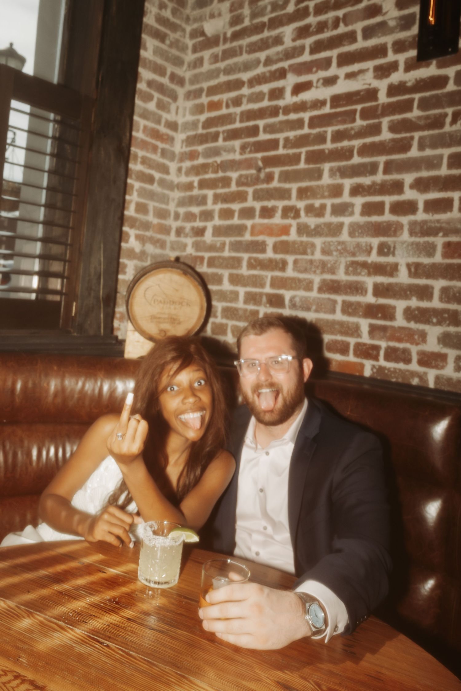 Couple smiling and making funny faces at a bar booth with brick wall background, holding cocktails.