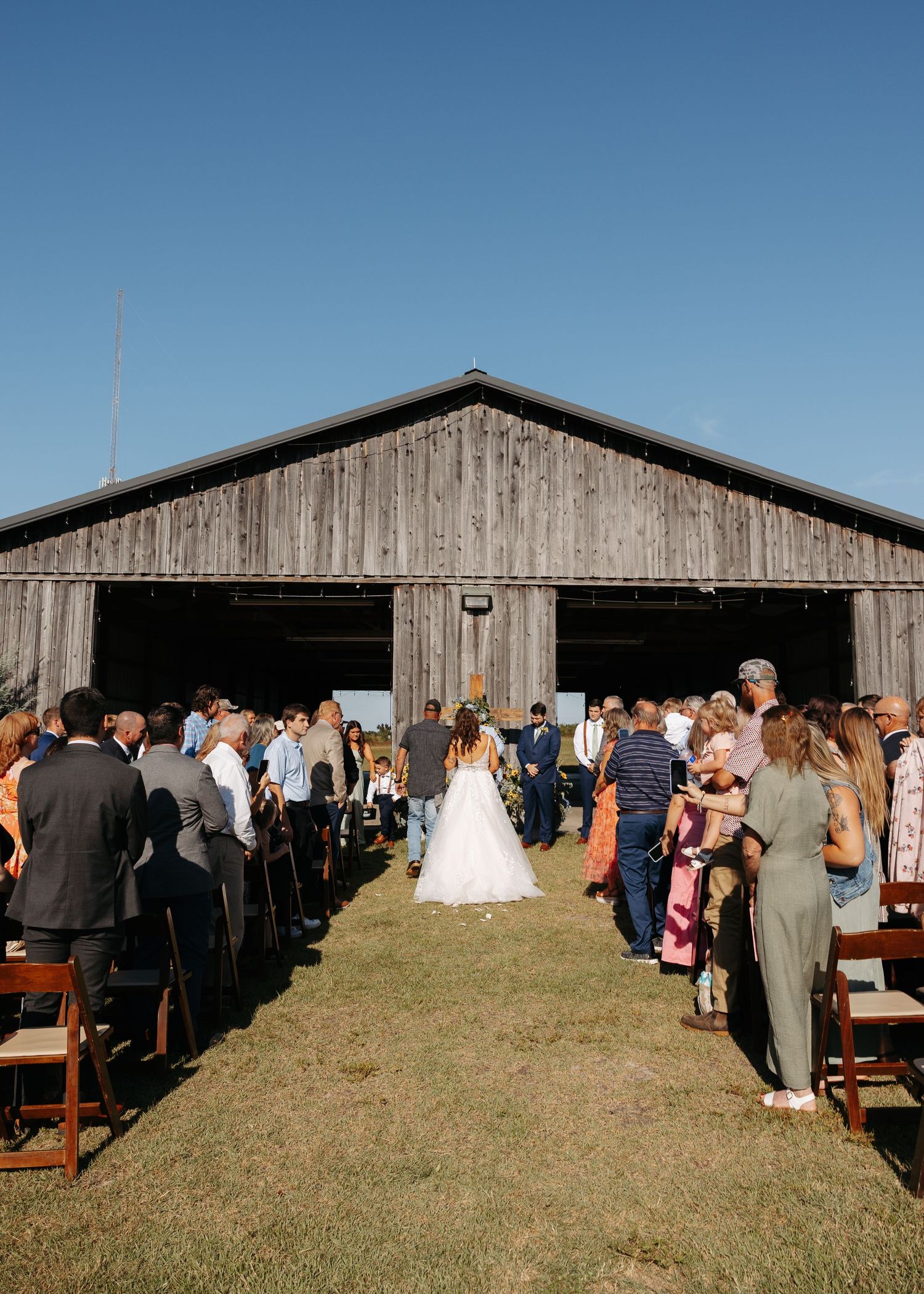 Bride walks down outdoor aisle toward rustic barn venue surrounded by wedding guests.