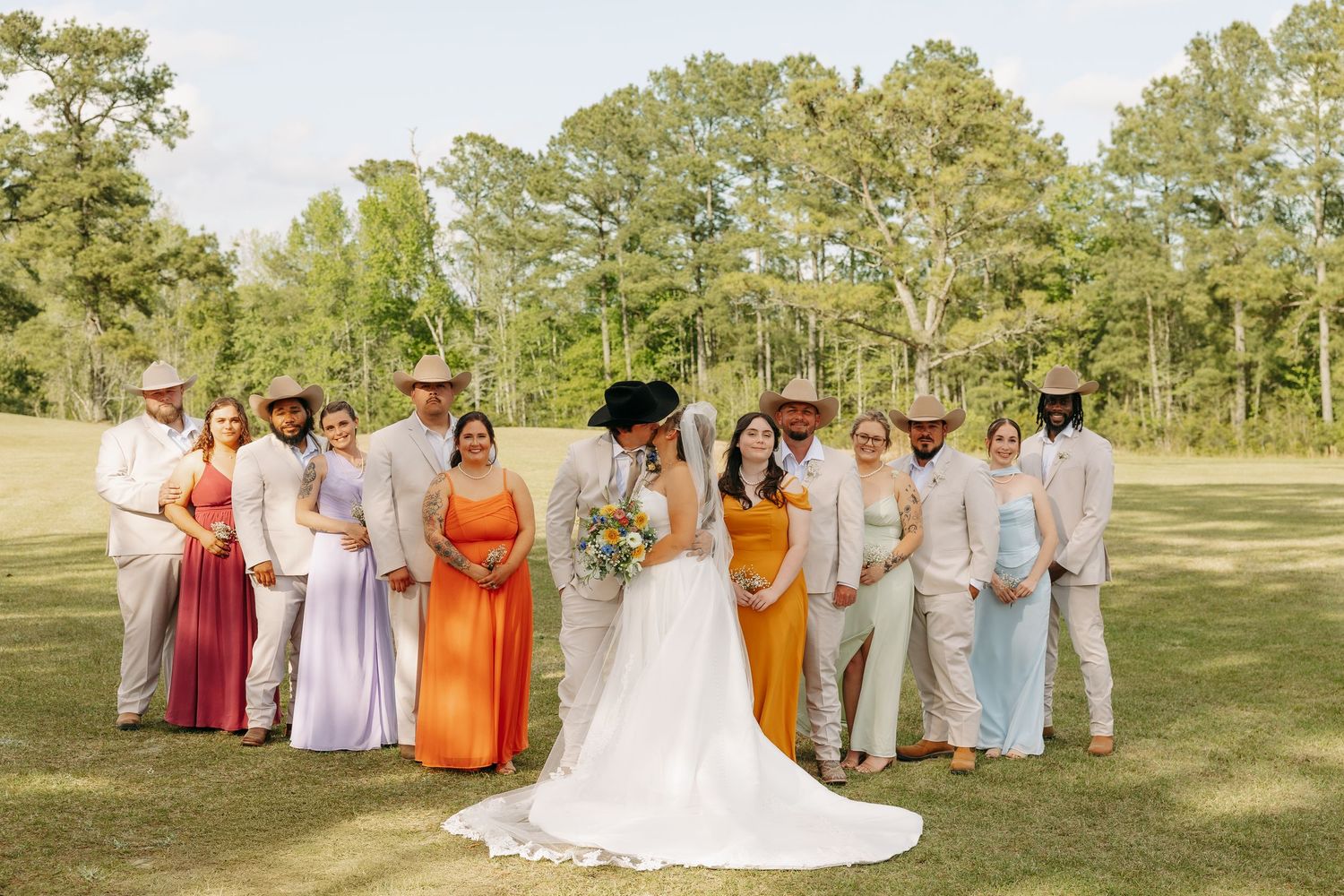 Wedding party posing outdoors on green lawn with bride, groom, groomsmen in tan suits and colorful bridesmaids.