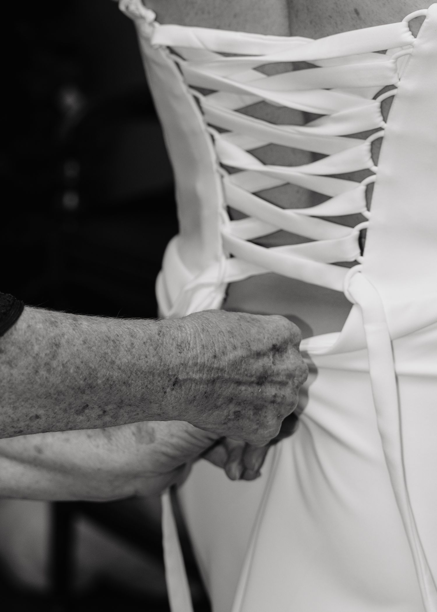 Elderly hands lacing up the back of a white wedding dress corset in a black and white photo.