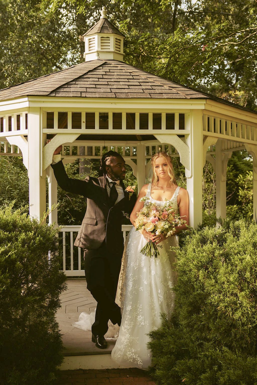 Bride and groom celebrating at a white garden gazebo, bride holding a bouquet, surrounded by lush greenery.