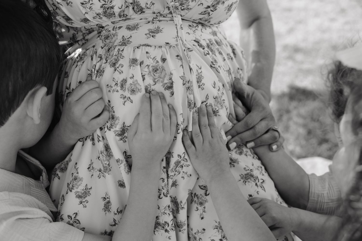 Black and white photo of a pregnant woman in floral dress with children's hands touching her baby bump.