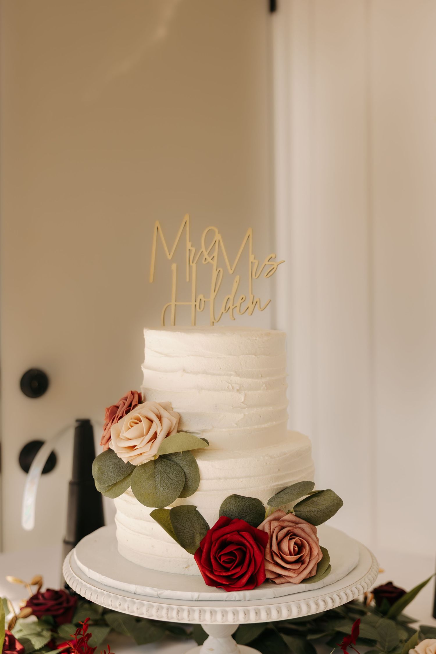 Elegant two-tier white wedding cake with Mr. & Mrs. Holden gold topper, adorned with red and pink roses and greenery.
