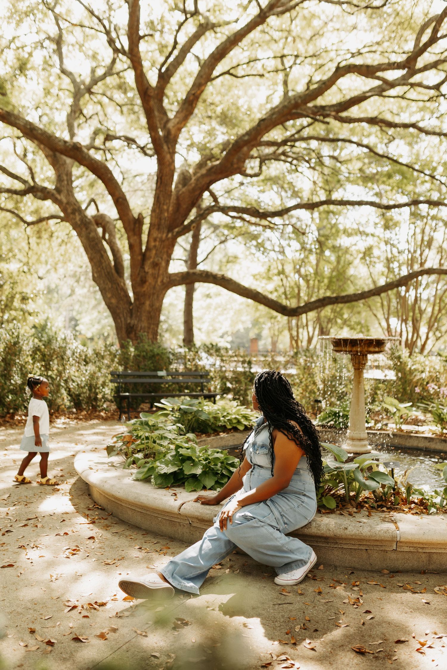 Woman in denim jumpsuit sits by a garden fountain as a child walks nearby under a large oak tree.