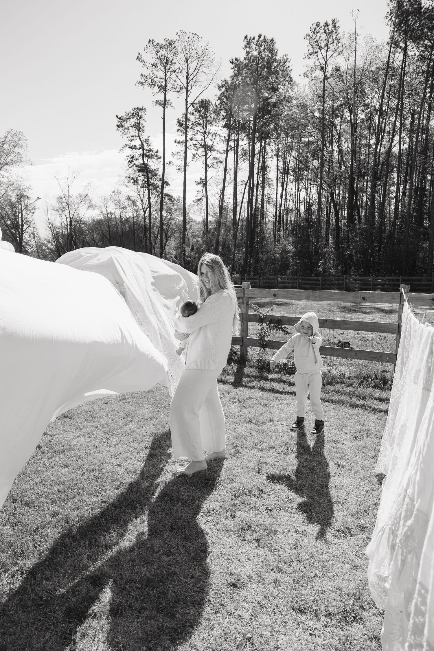 Black and white photo of a woman and child hanging white sheets on a clothesline outdoors near a wooden fence.