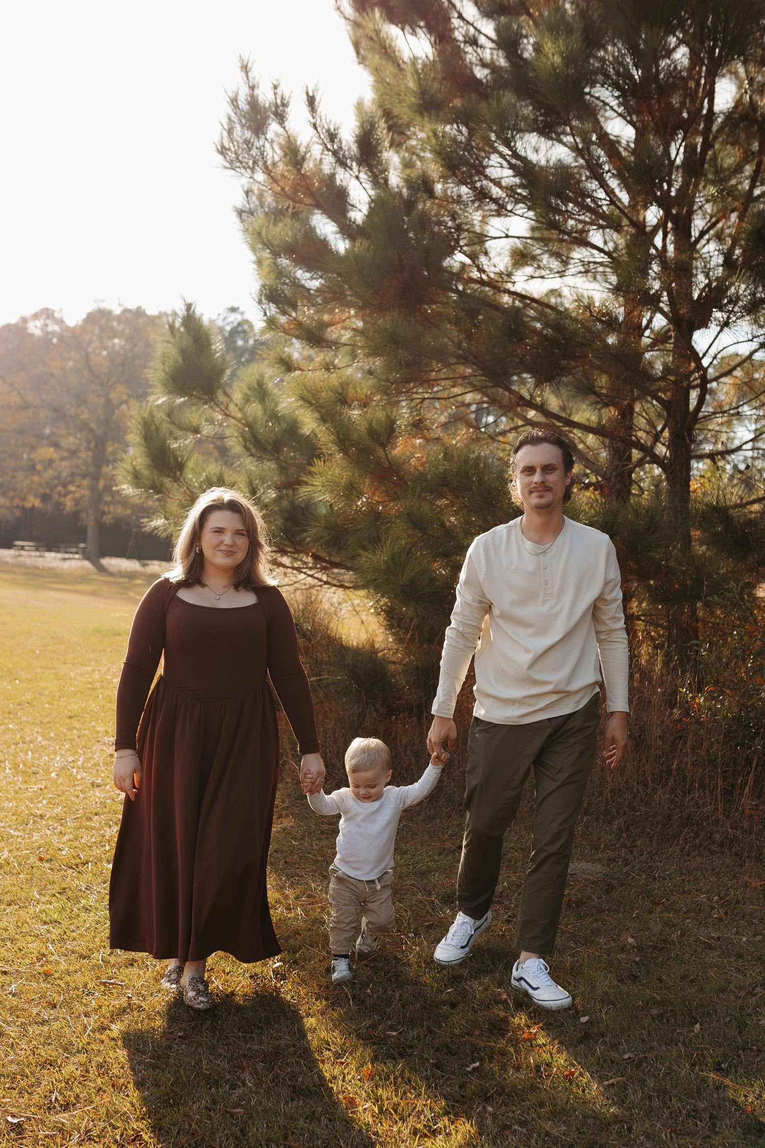 Family of three walking outdoors in autumn, parents holding toddler's hands near pine trees in golden sunlight.