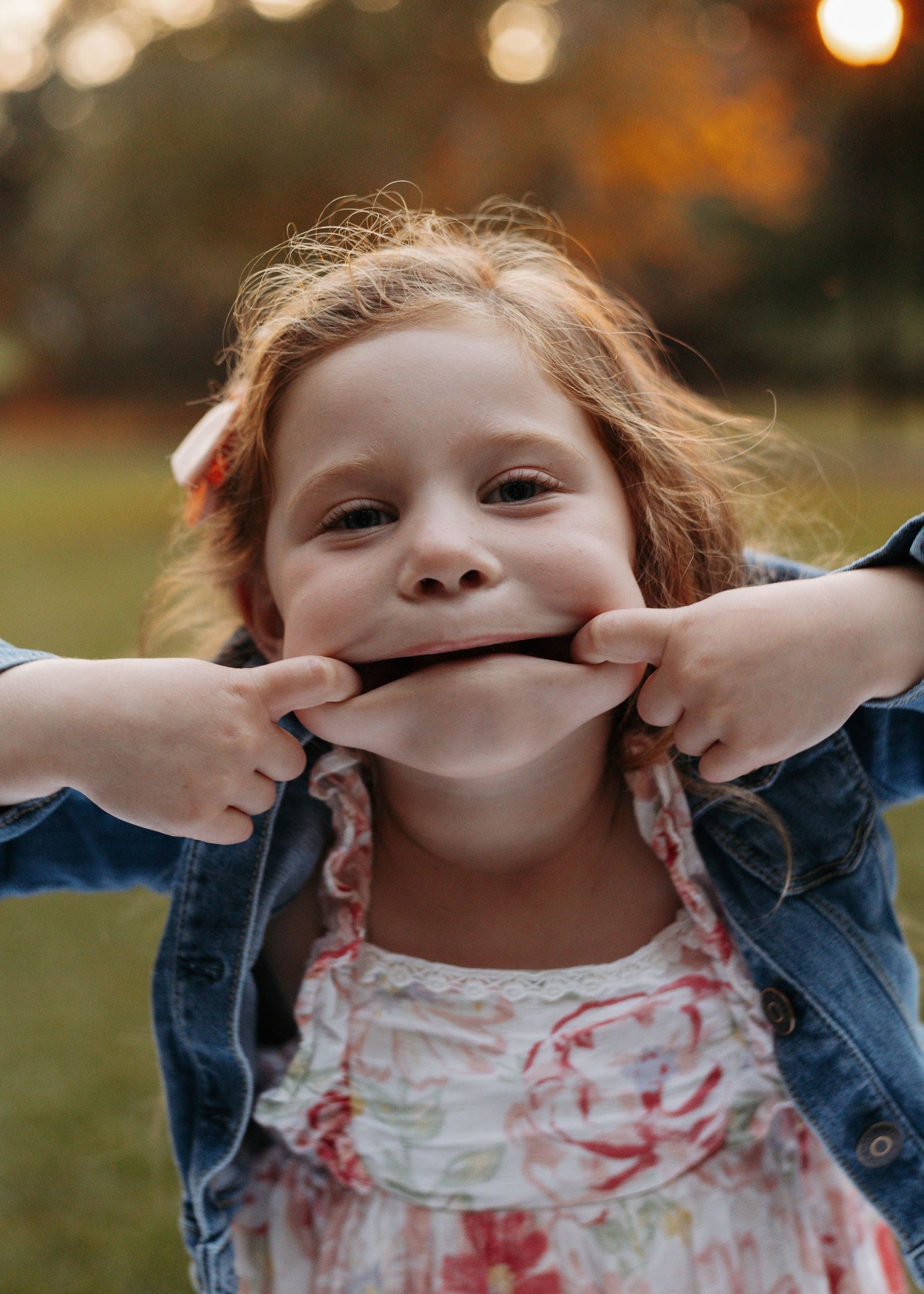 A playful young girl with red hair stretches her smile wide with her fingers outdoors in warm golden light.