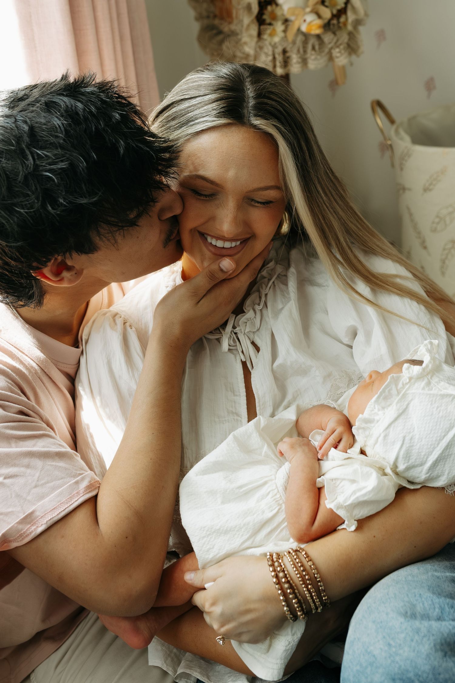 A loving couple shares a tender kiss while cradling their newborn baby in a soft, warmly lit nursery.