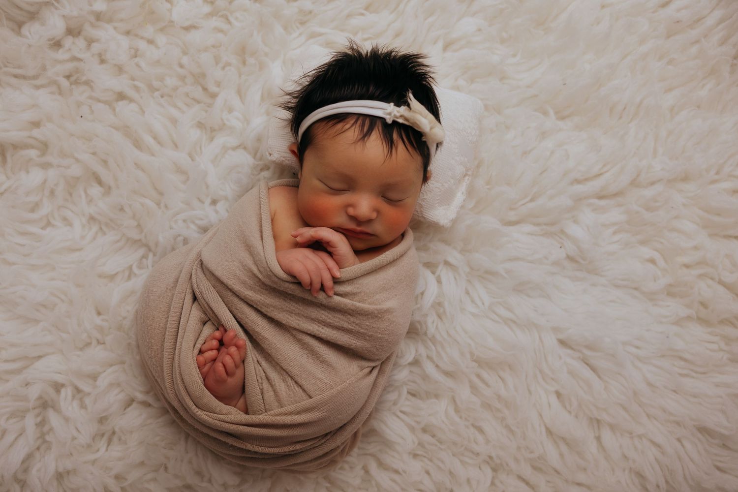 Newborn baby wrapped in beige blanket sleeping peacefully on soft white textured backdrop.