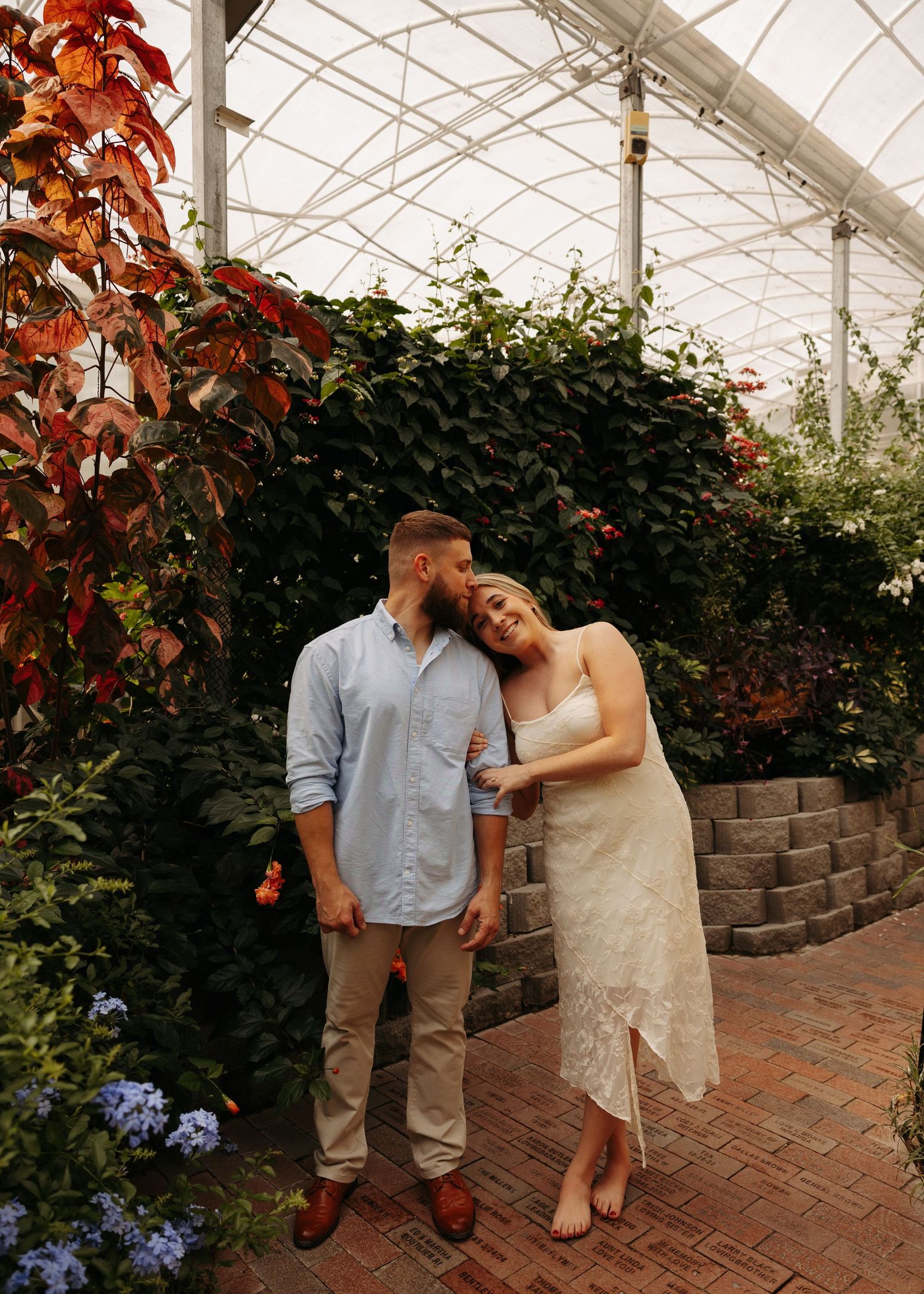 Couple embracing in a lush greenhouse, woman in lace dress leans on man in blue shirt amid colorful foliage.