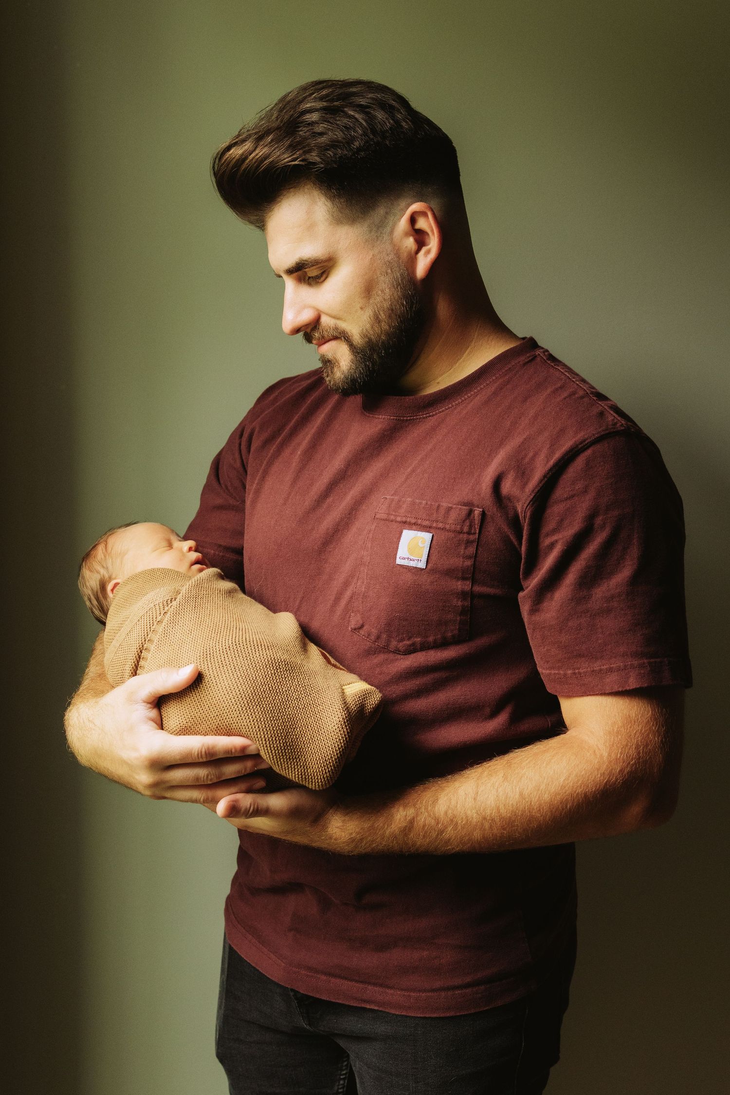 A person in a burgundy shirt cradles a tiny bundle wrapped in a brown blanket in a dimly lit studio setting.