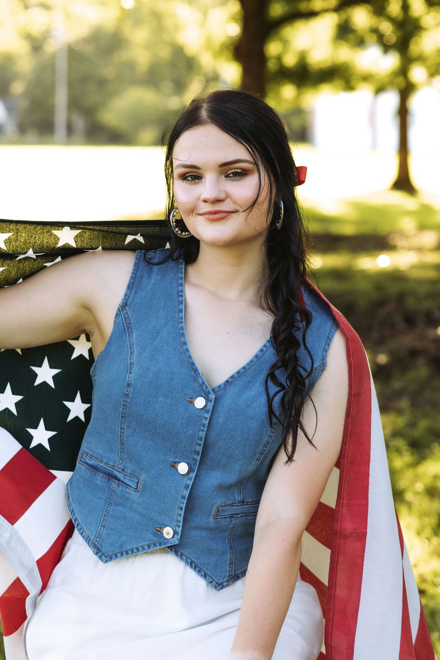 Person wearing denim vest and white dress holding American flag in sunny outdoor setting.