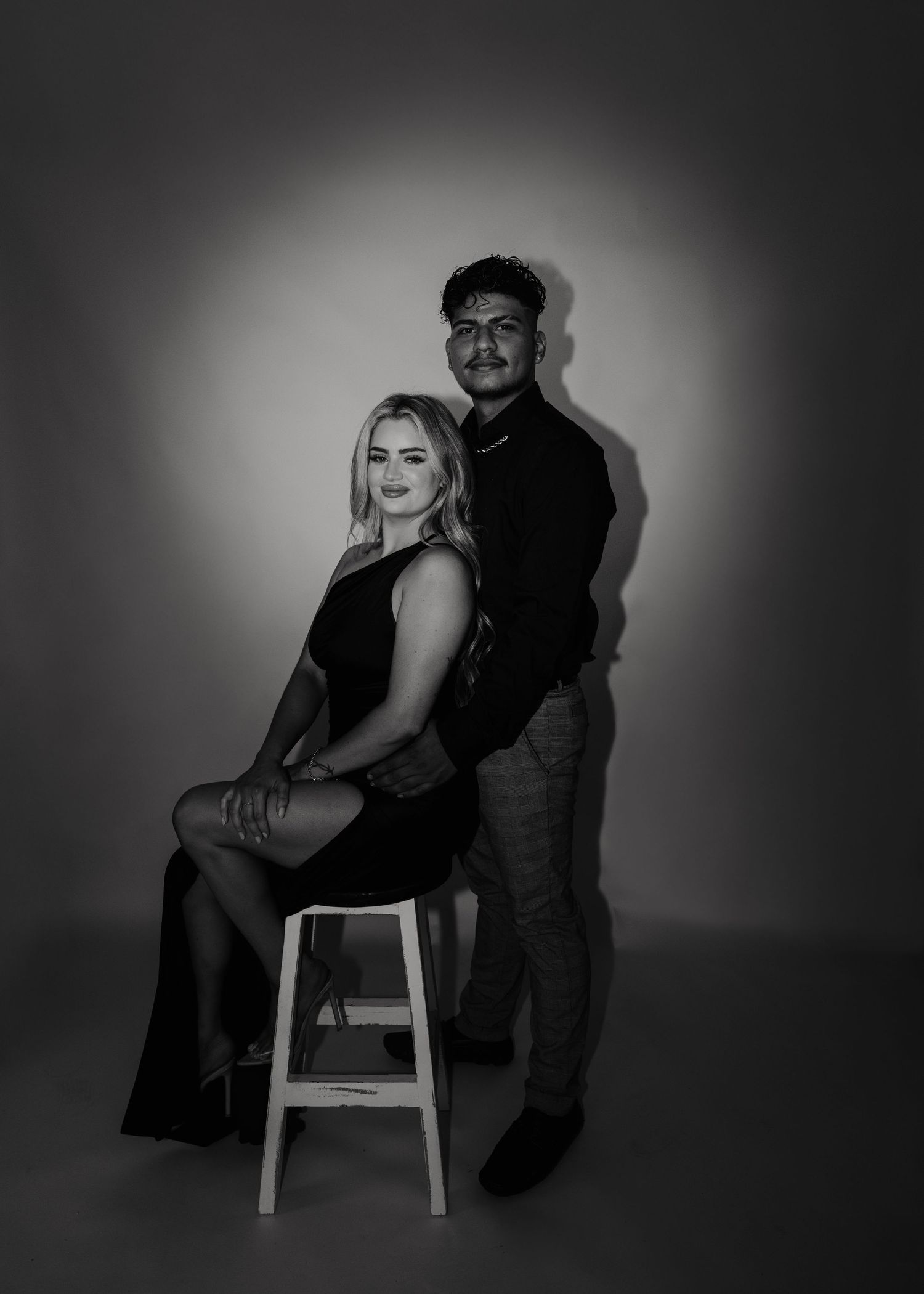Black and white studio portrait of a couple posing together, with one person seated on a stool and another standing behind.