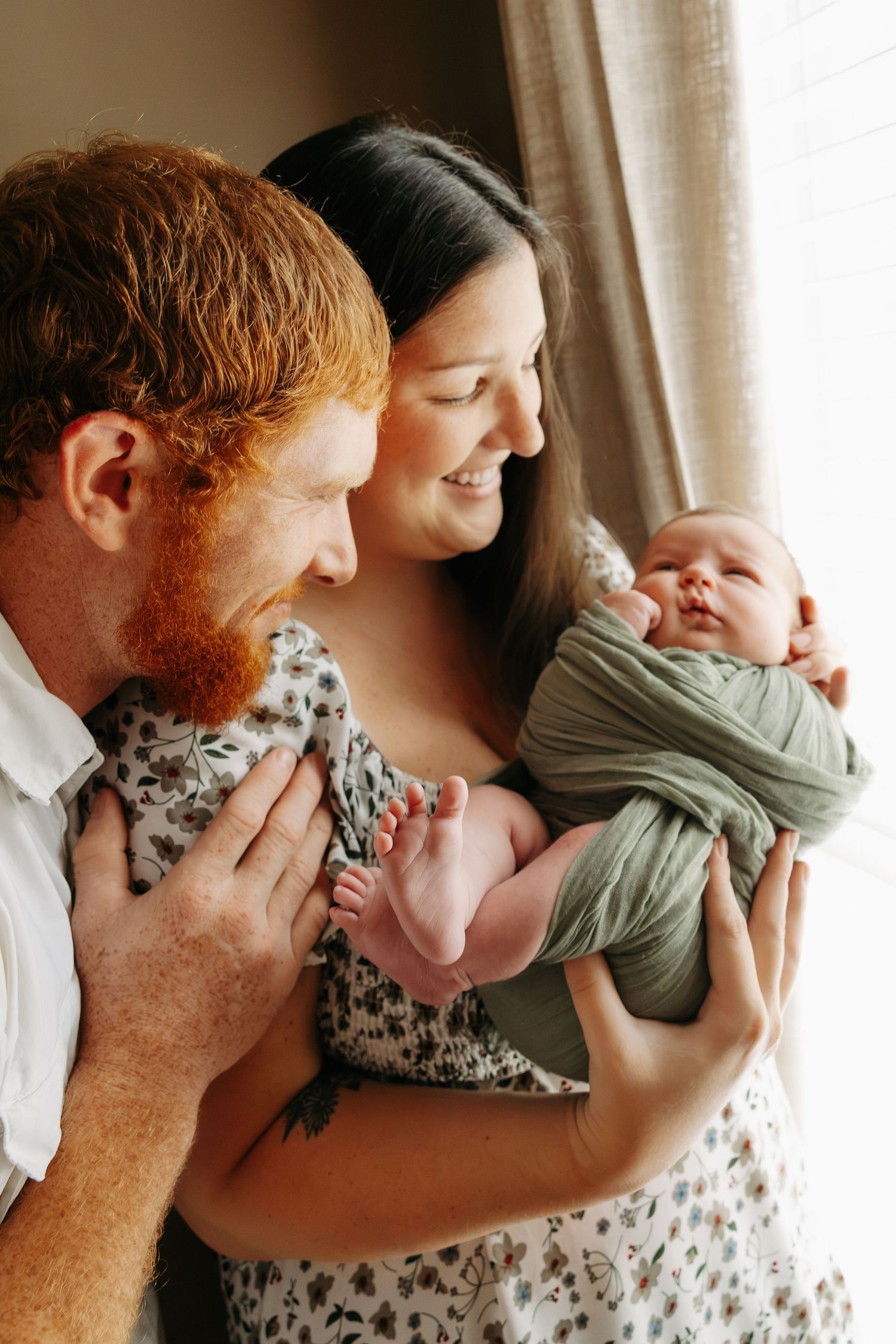 Happy parents with red-haired father and dark-haired mother smiling at their newborn baby wrapped in a green swaddle.