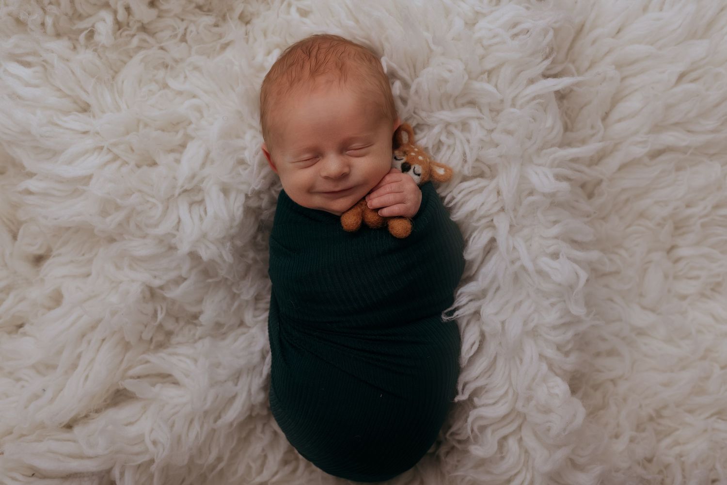 Smiling newborn baby swaddled in dark green wrap lying on fluffy white rug holding a small toy.