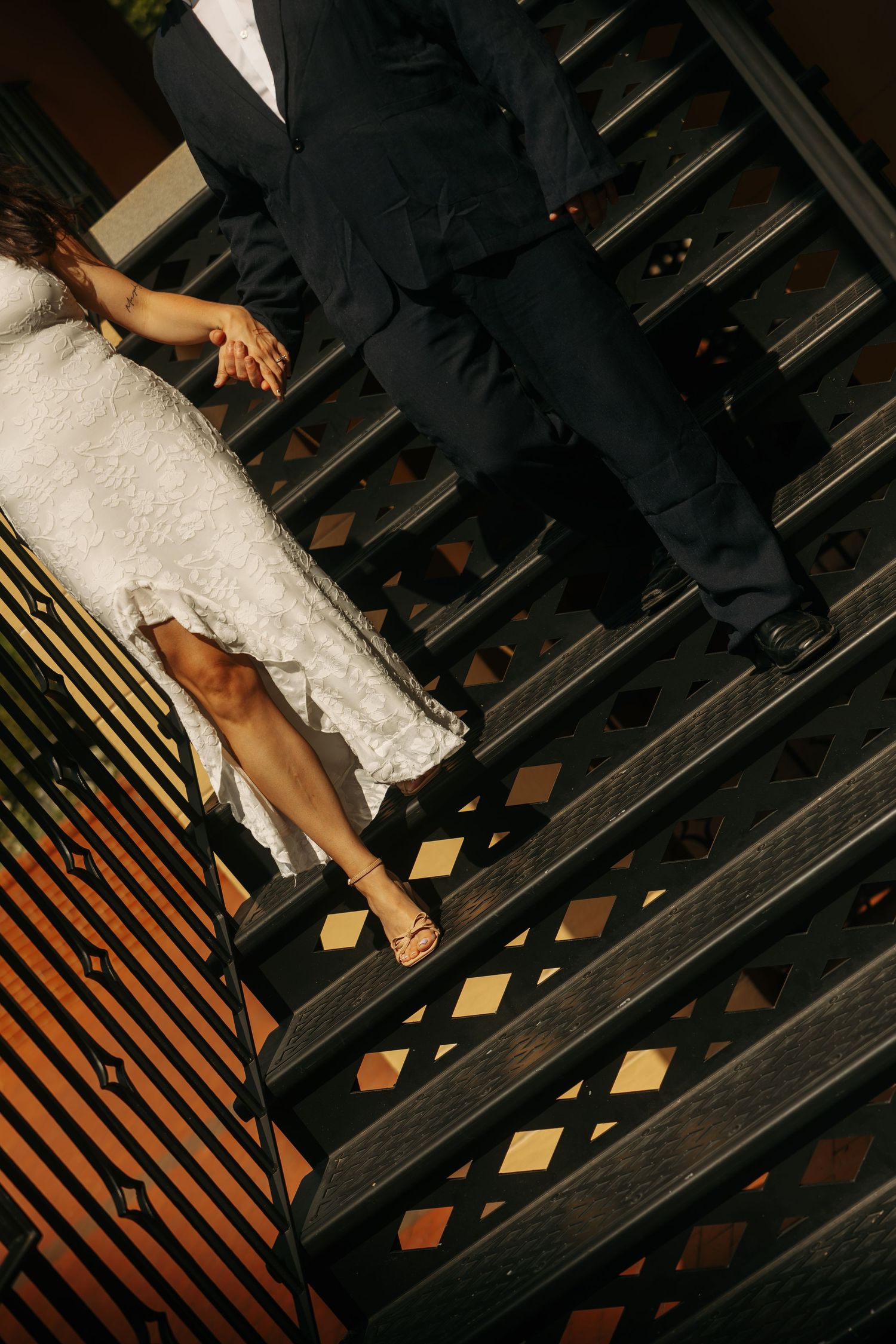 Bride in white lace dress and groom in black suit holding hands while descending elegant dark wooden staircase.