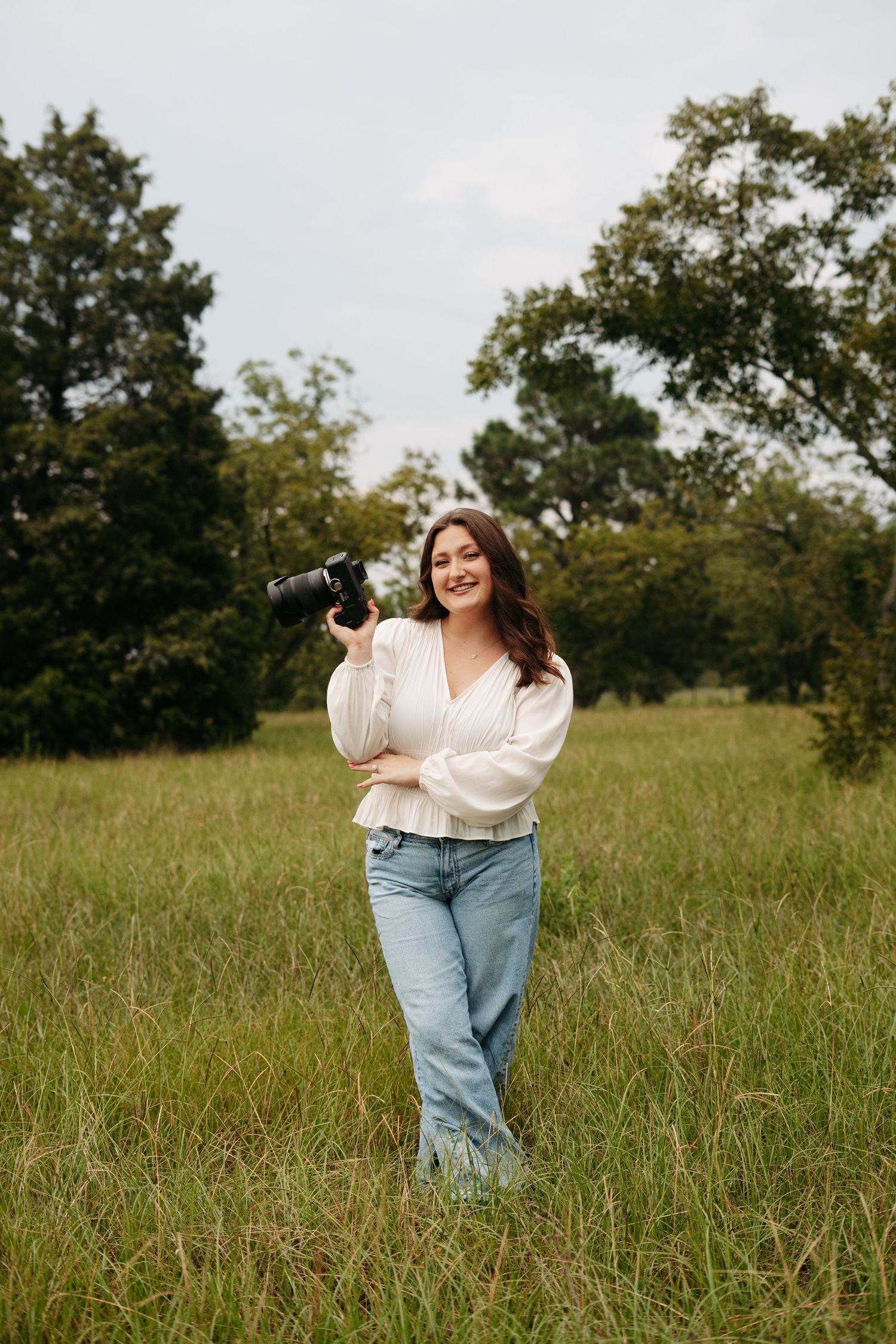 A photographer wearing a cream sweater and light jeans stands in a grassy field on a sunny day.