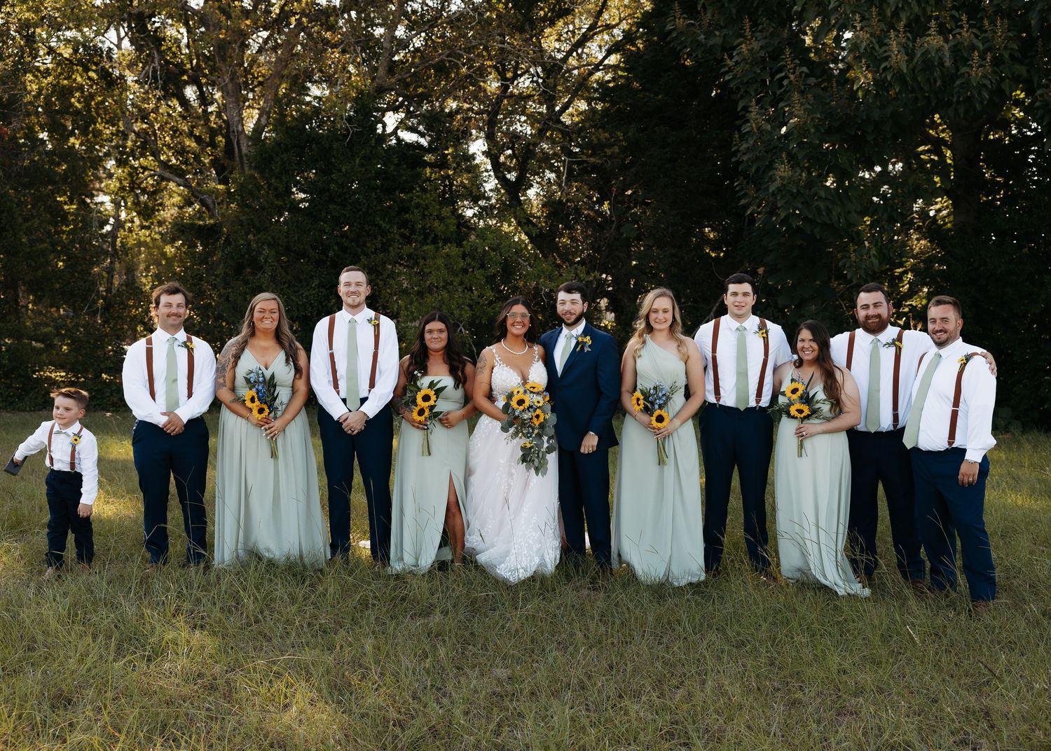Wedding party posing outdoors in a grassy field, bridesmaids in sage green gowns, groomsmen in navy suits with suspenders.