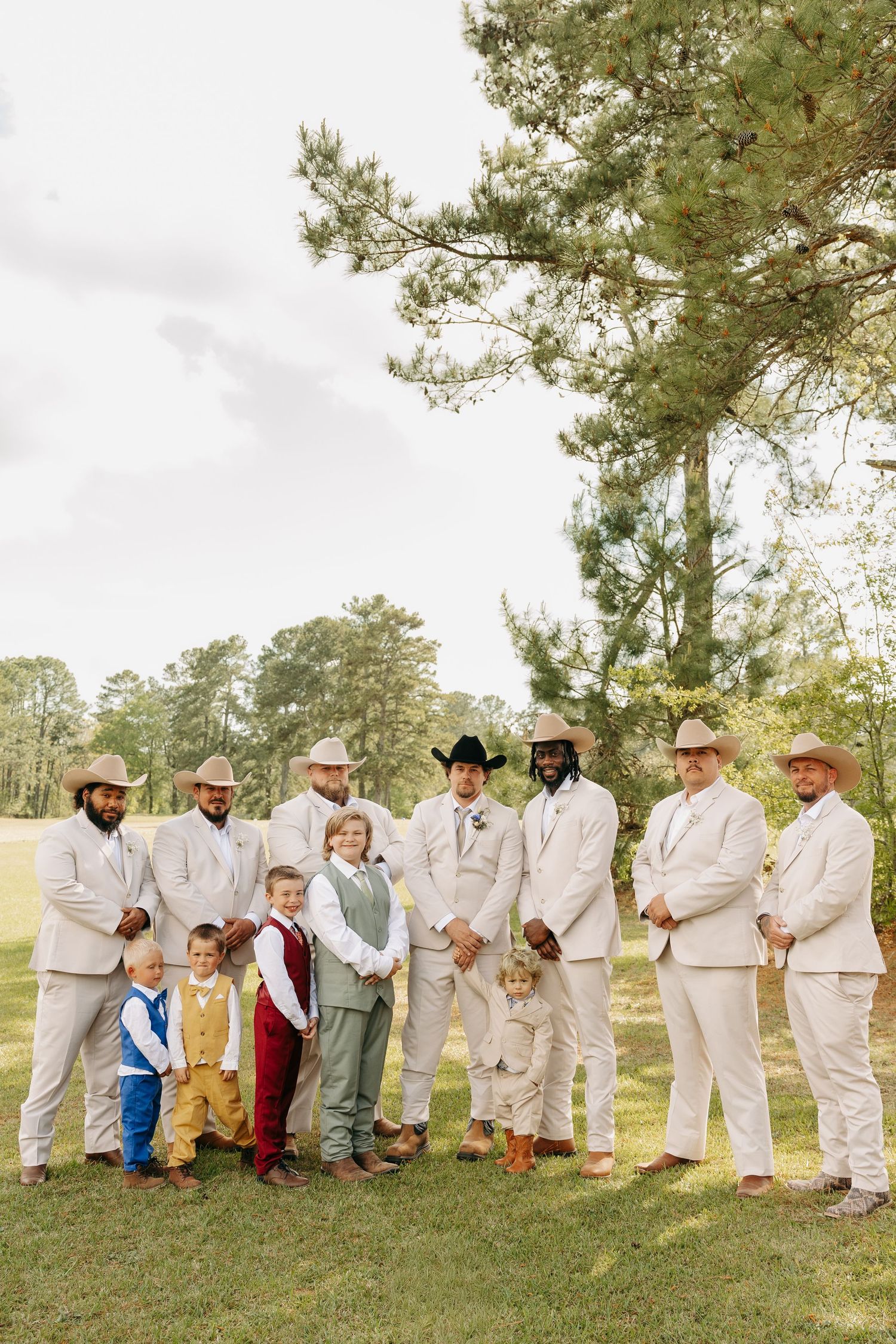 Groomsmen in tan suits and cowboy hats pose outdoors with young boys in colorful outfits at a country wedding.