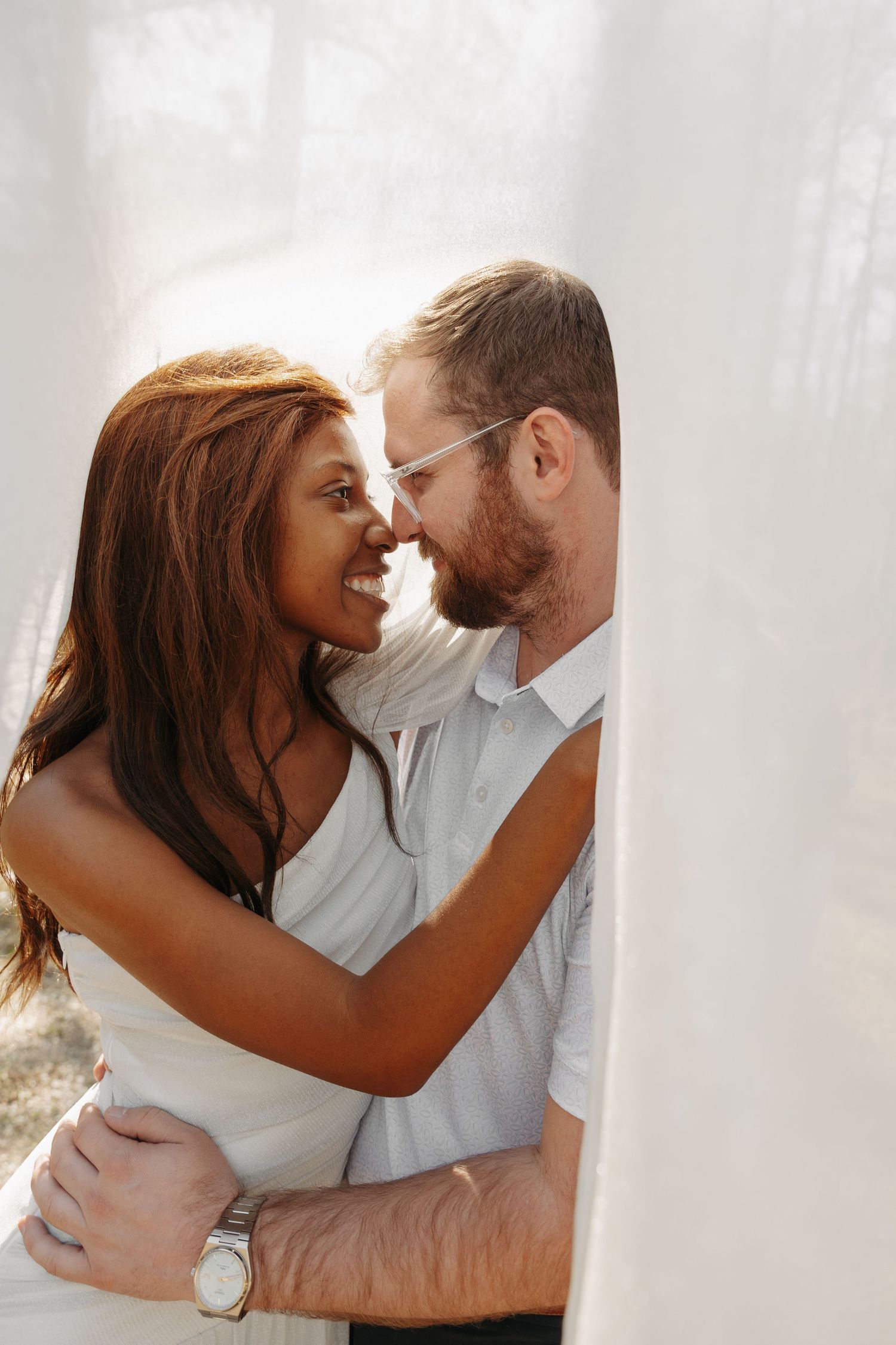 Interracial couple sharing a romantic moment, nearly kissing, embraced in soft white light outdoors.