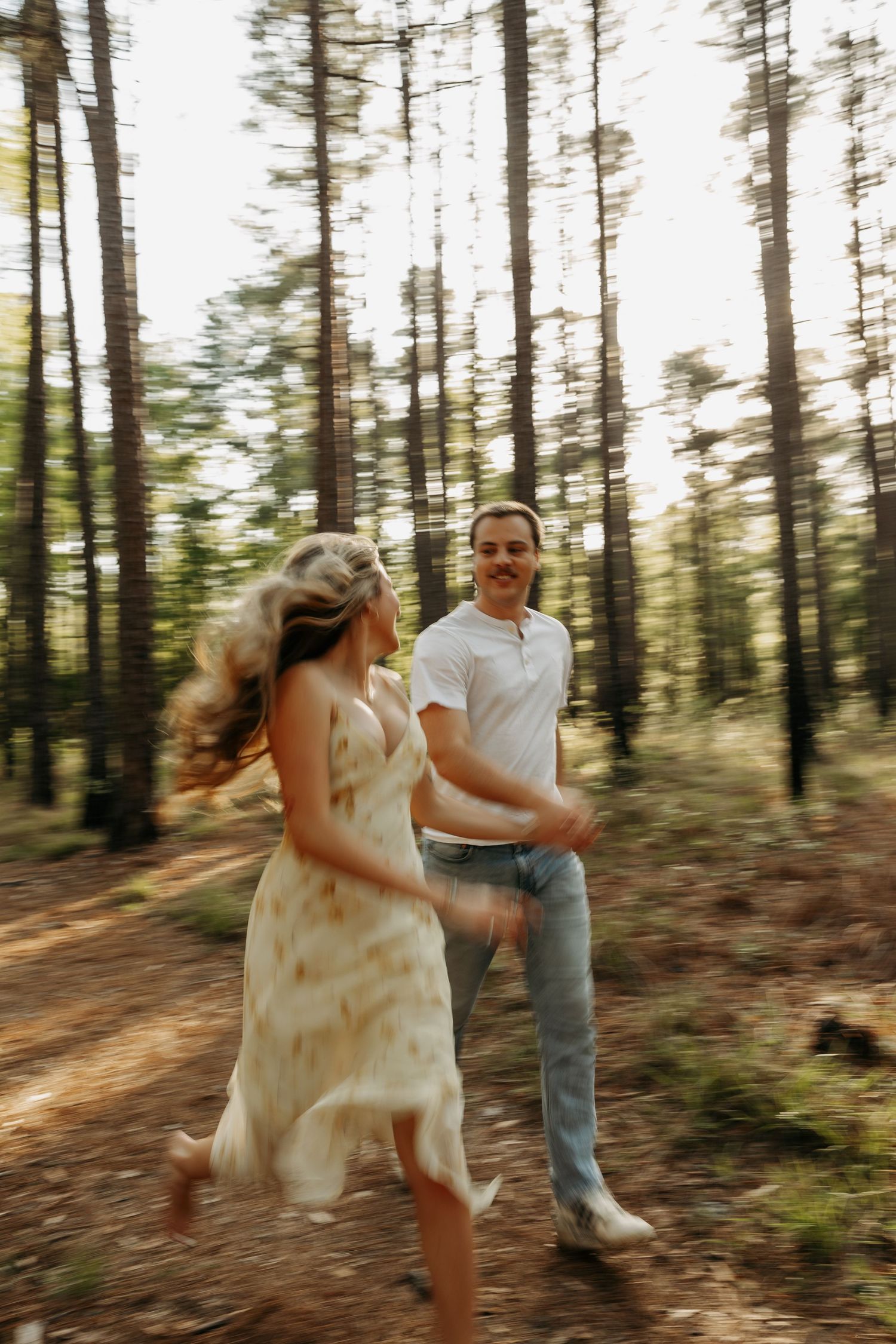 A couple joyfully runs through a sunlit pine forest, the woman in a floral dress, the man in a white tee and jeans.