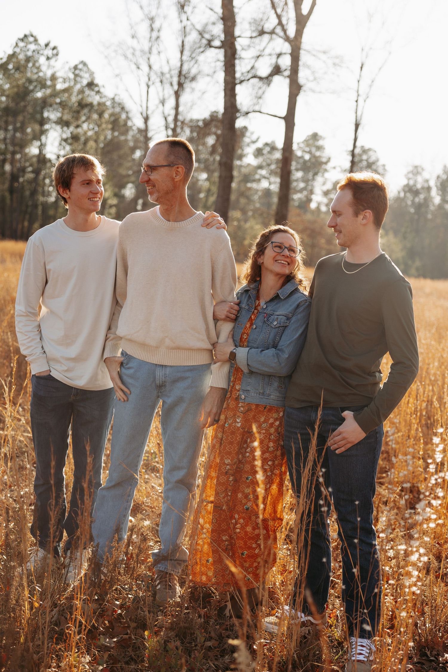 Family of four smiling and walking together in a sunlit autumn field surrounded by tall golden grass and pine trees.