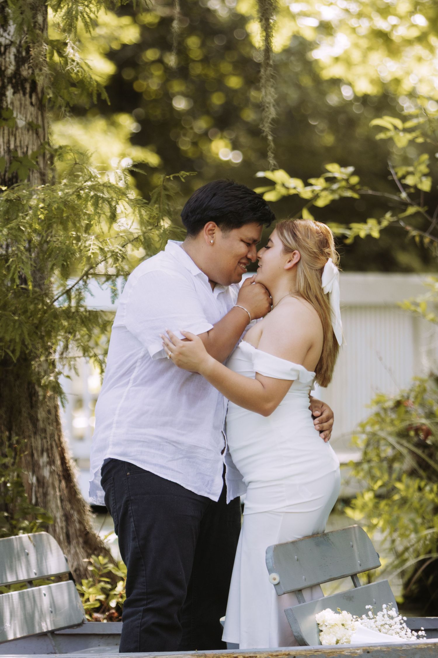 Couple in white outfits sharing a romantic moment outdoors, surrounded by lush green trees.