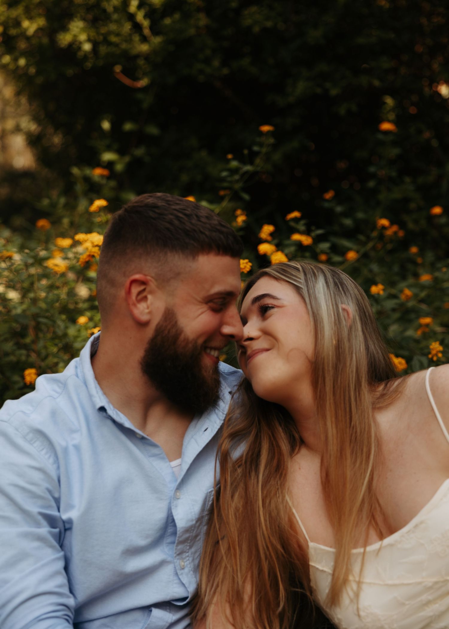 A smiling couple sharing a romantic moment outdoors surrounded by yellow flowers in a lush garden.