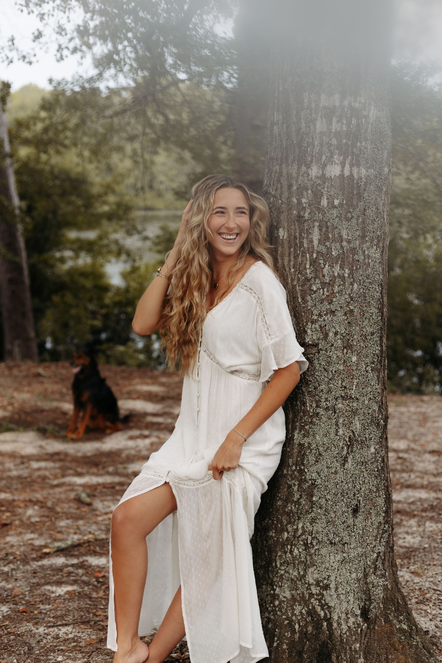 A person in a flowing white dress poses playfully against a tree trunk in a dreamy outdoor setting.
