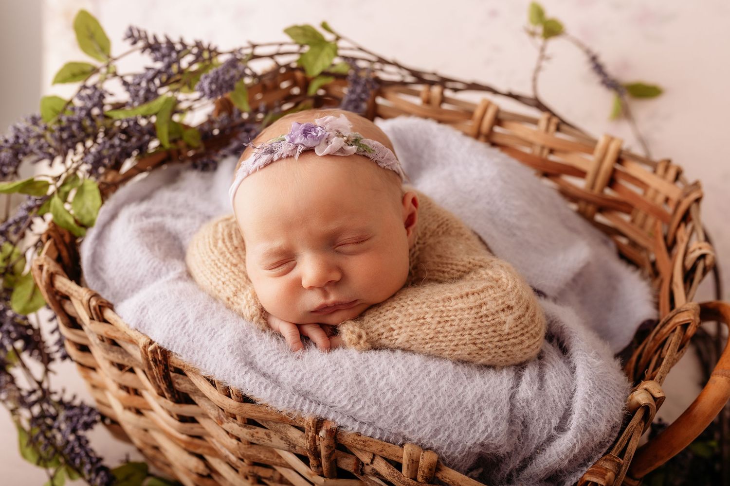 Newborn baby sleeping peacefully in a wicker basket with lavender flowers and soft blue blanket.