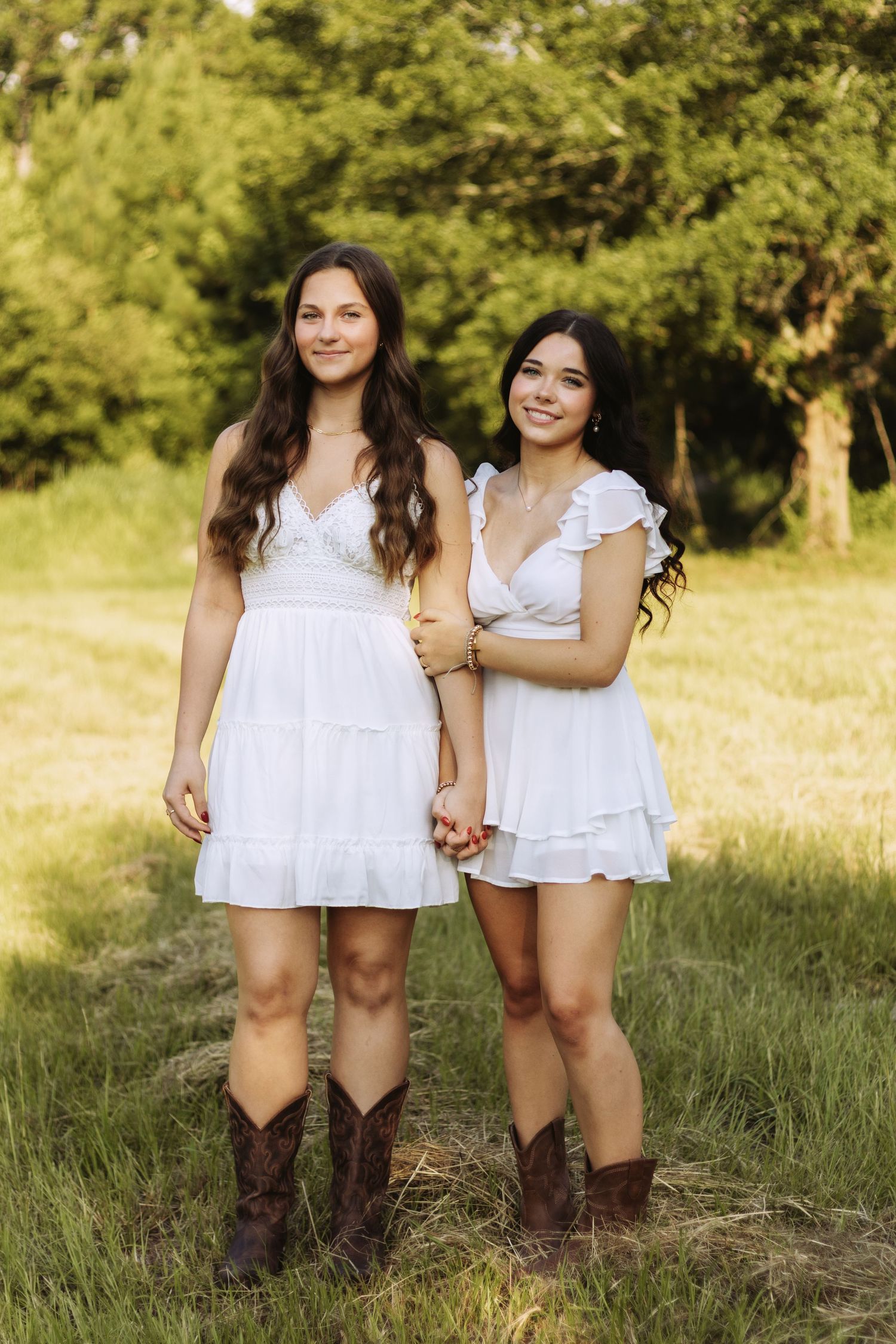 Two people in white summer dresses and brown cowboy boots pose together in a sunny grassy field with trees in background.