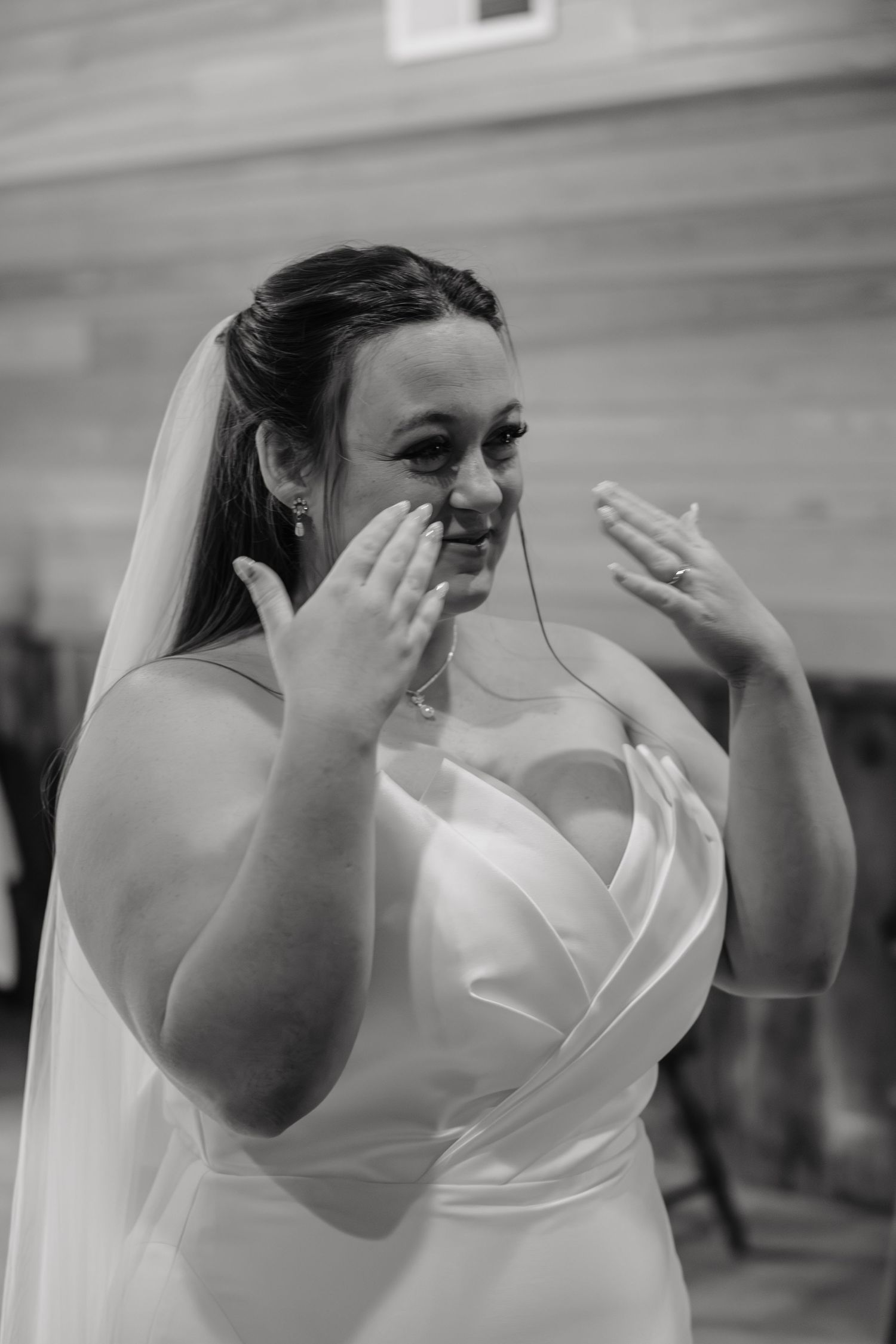 Emotional bride in white strapless gown and veil wipes tears during wedding ceremony in black and white photo.