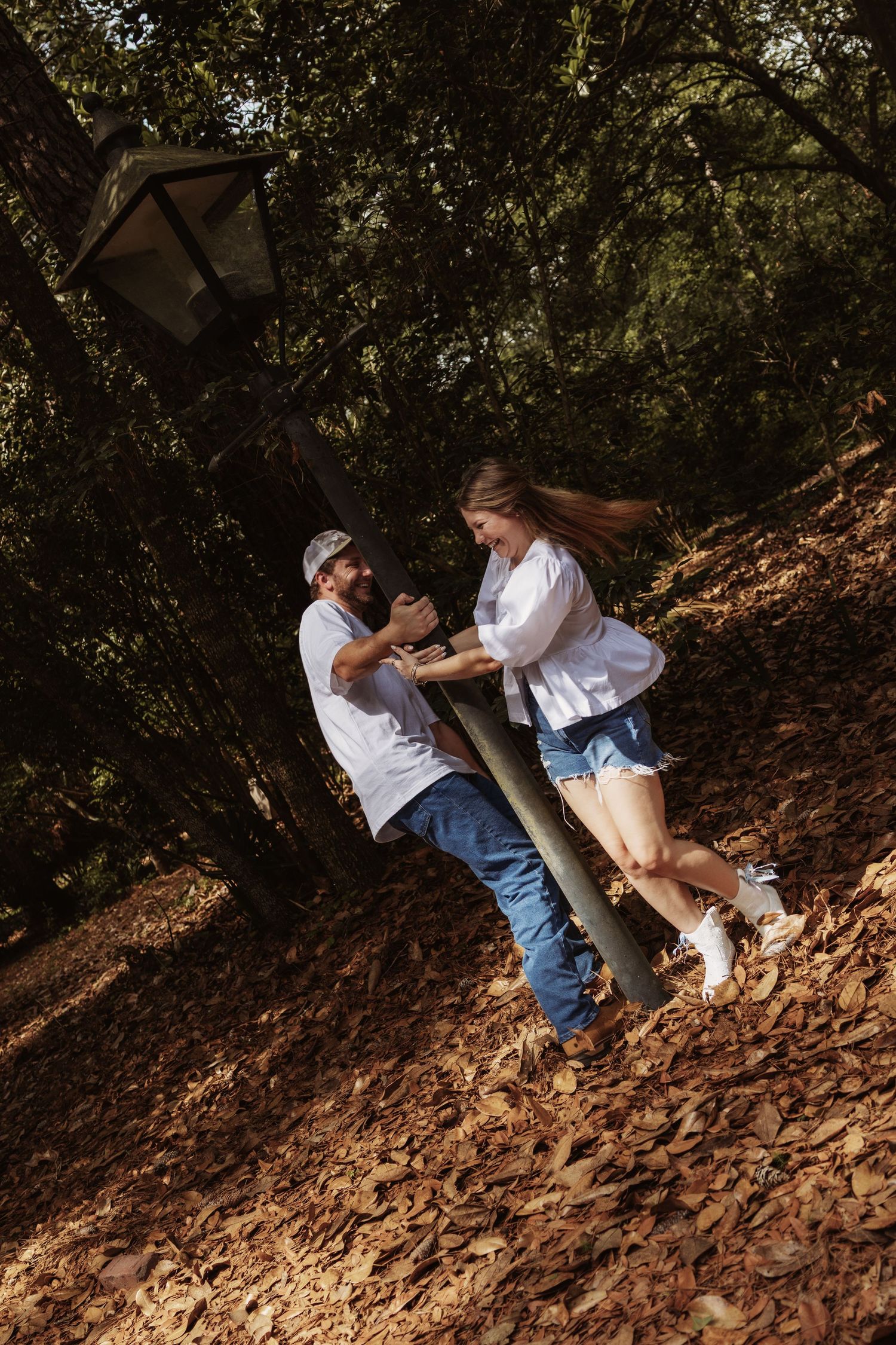 Couple playfully spinning around a lamppost on a leaf-covered forest path in autumn sunlight.