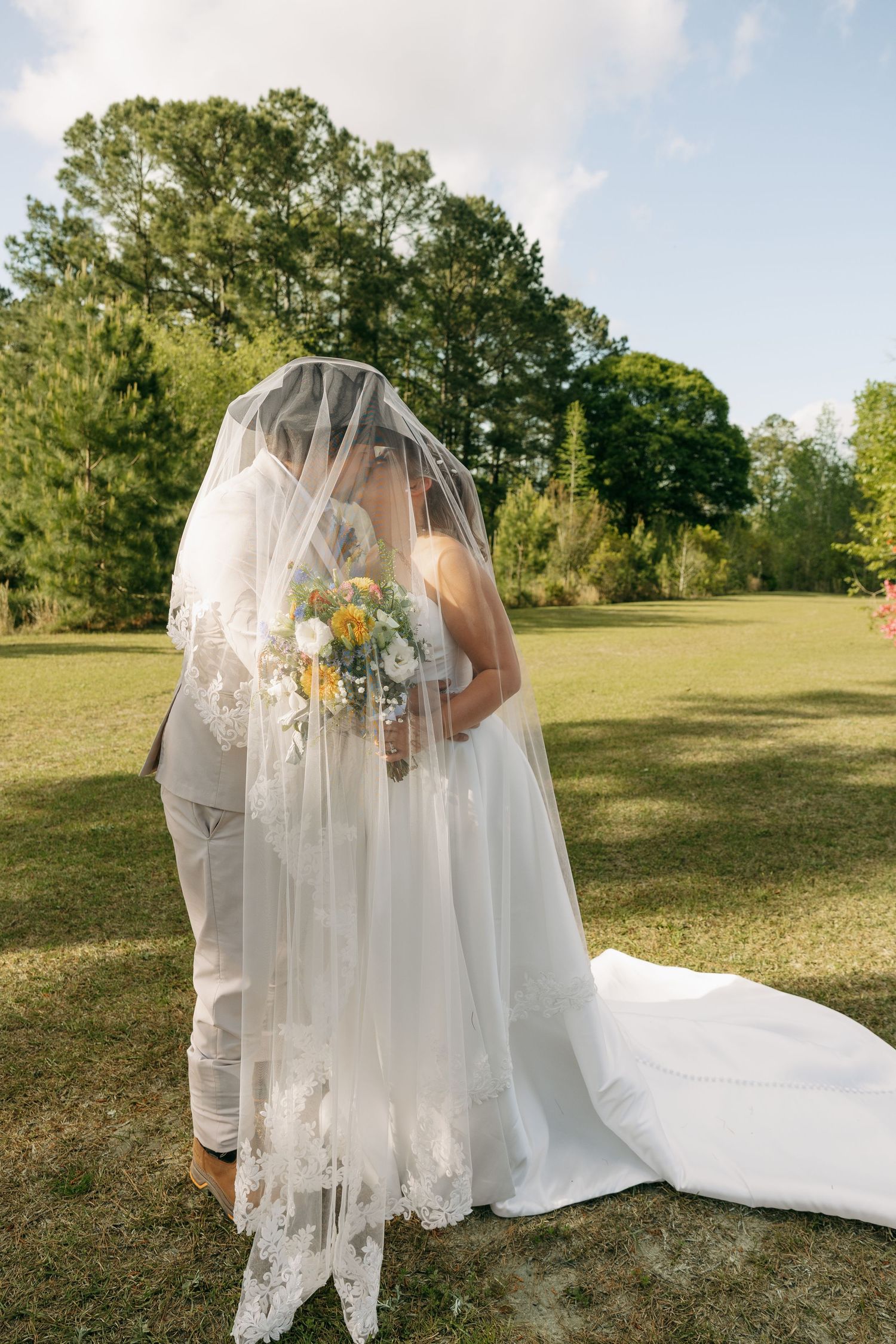 Bride and groom kissing under a lace veil outdoors, bride holding a yellow and white bouquet.