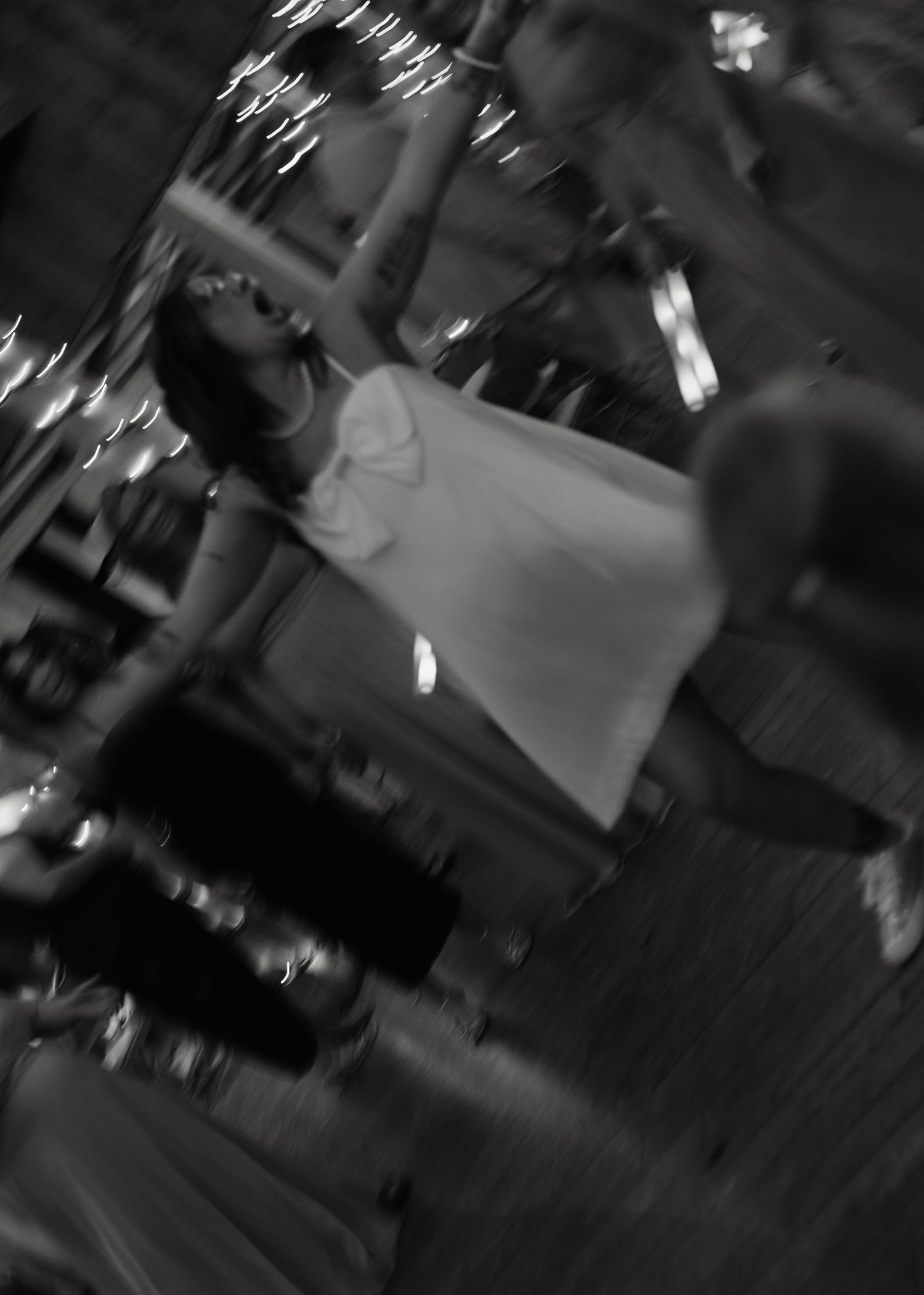 Blurry black and white photo of a woman in a white dress dancing at a dimly lit event.