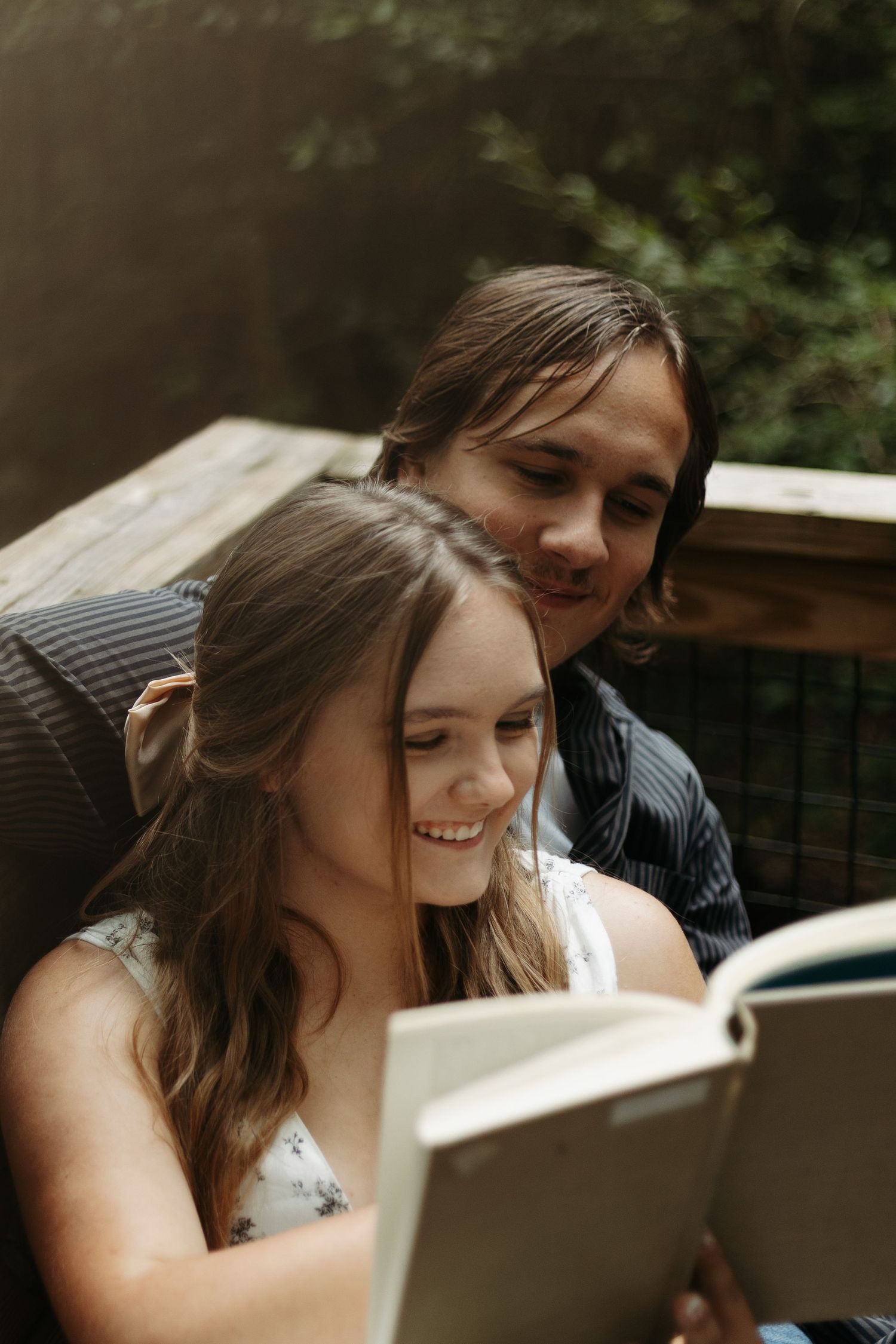 A young couple smiling and reading a book together outdoors on a wooden deck surrounded by greenery.