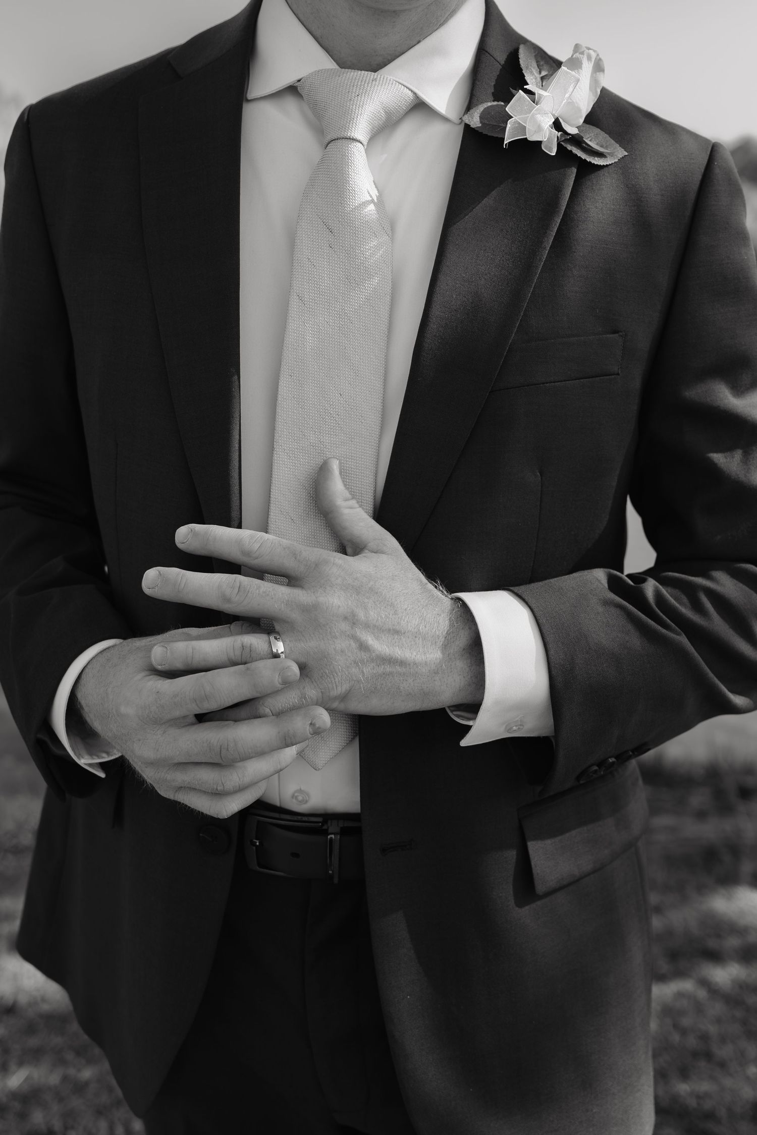 Black and white close-up of groom in dark suit adjusting wedding ring with floral boutonniere.
