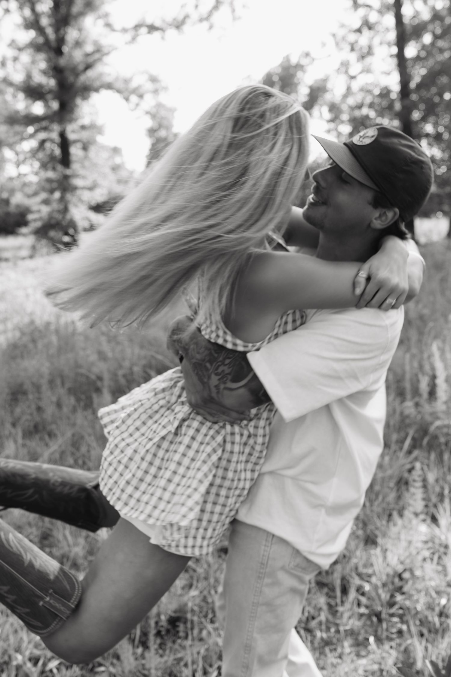 Black and white photo of a couple embracing outdoors, man lifting woman as her hair flows, surrounded by trees.