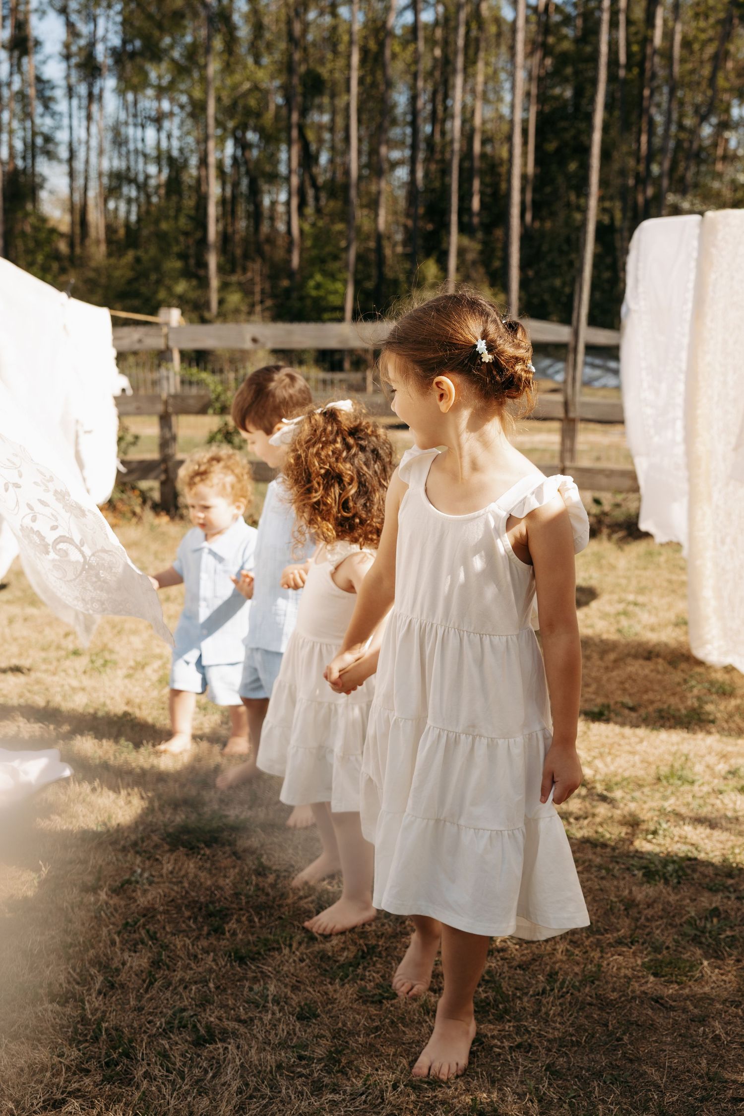 Children in white dresses play barefoot outdoors near hanging laundry on a sunny day at a rustic farm.