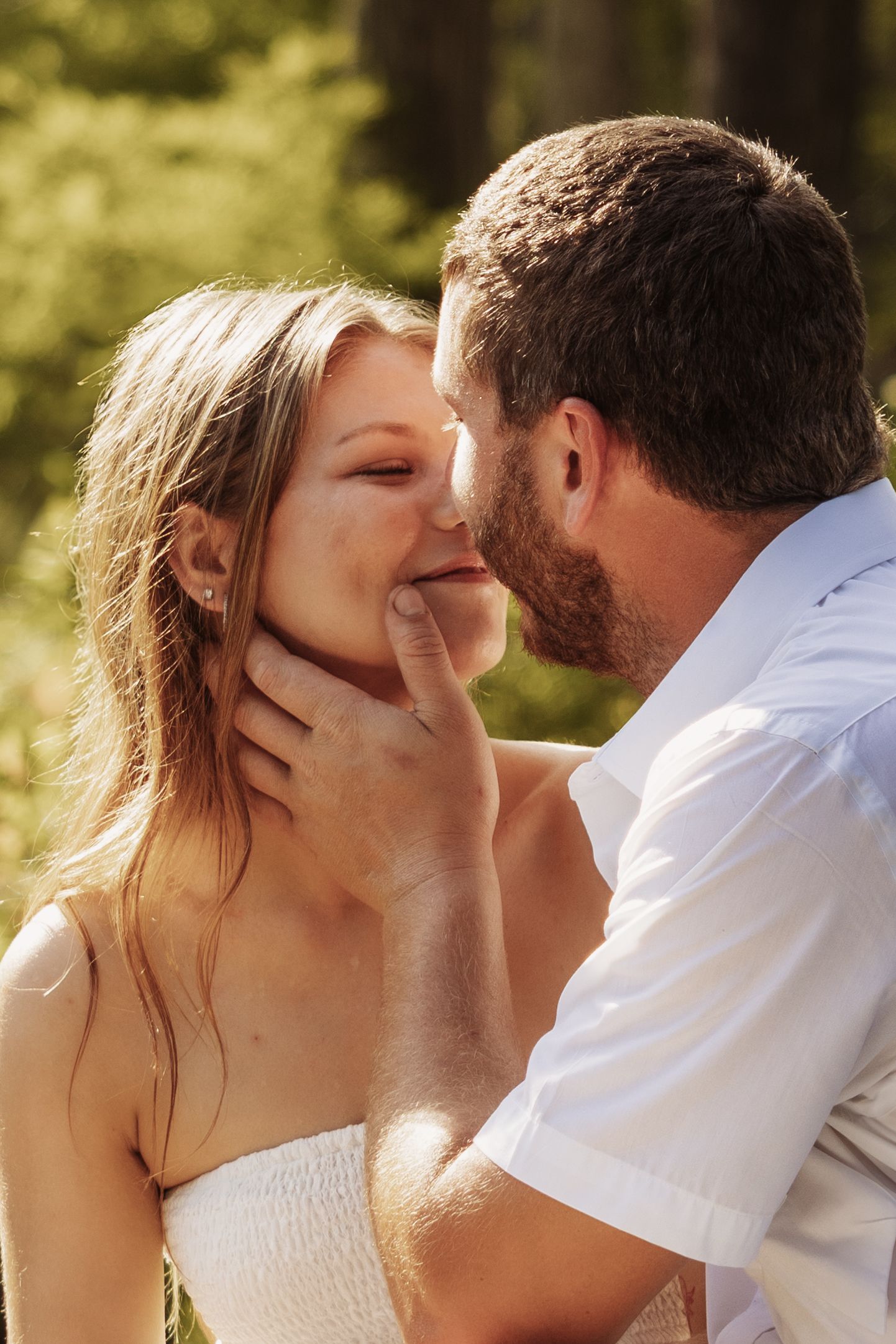 A bearded man in a white shirt gently holds a smiling woman's chin while leaning in for a kiss outdoors.
