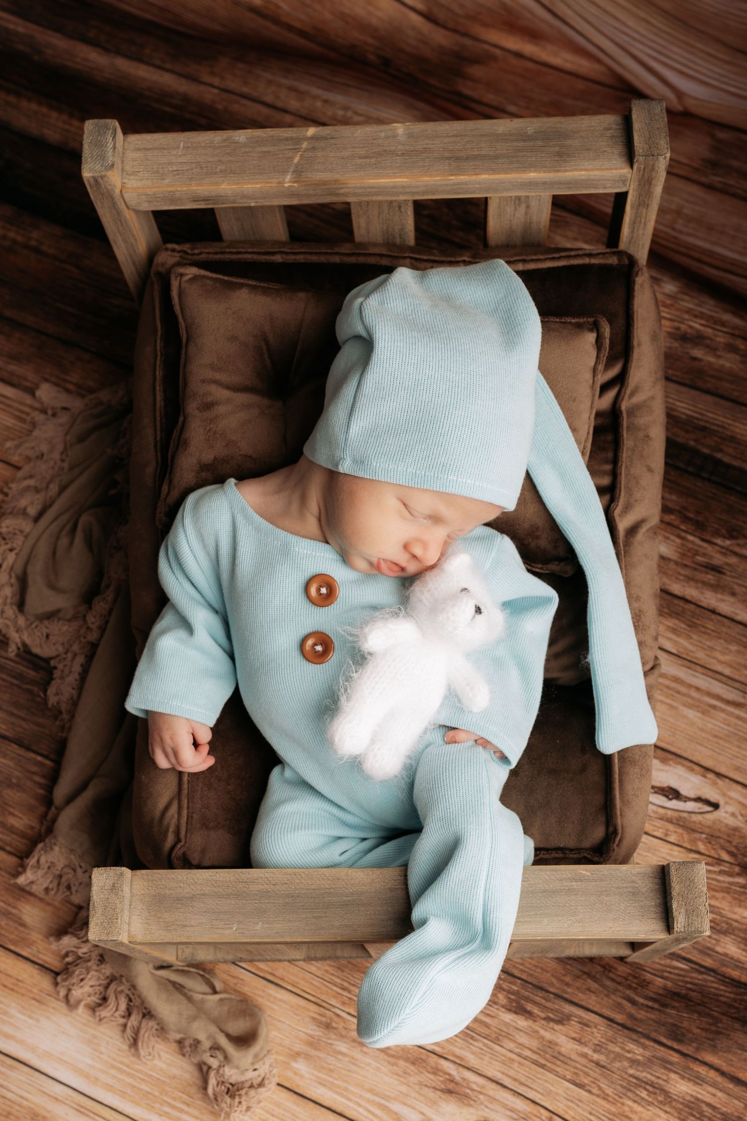 A sleeping baby wearing light blue pajamas and hat rests in a wooden box with brown cushions while holding a white plush toy.