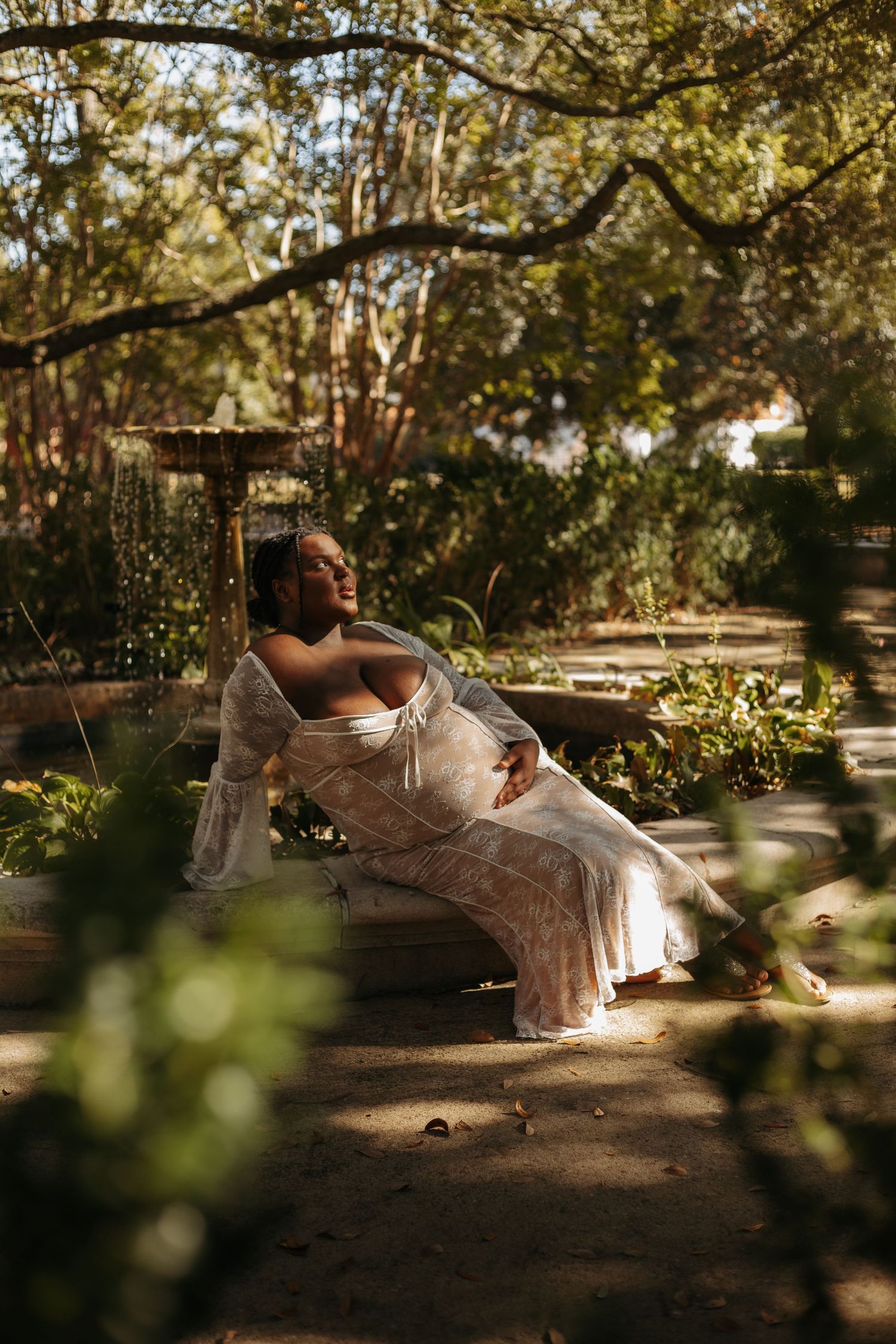 Pregnant woman in white lace dress relaxing on bench in sunlit garden surrounded by lush green trees.