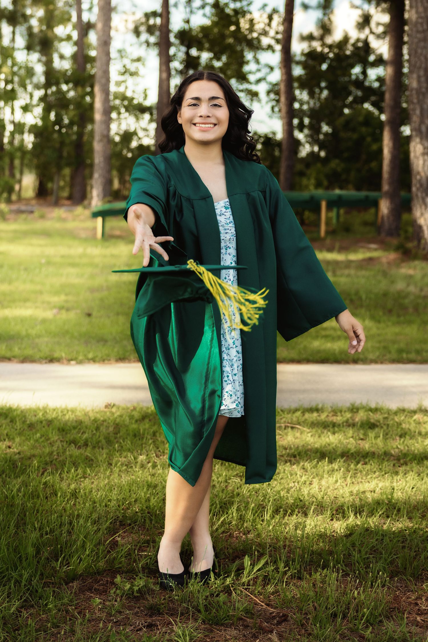 Graduate wearing floral dress and green gown sits gracefully on grass in outdoor setting.