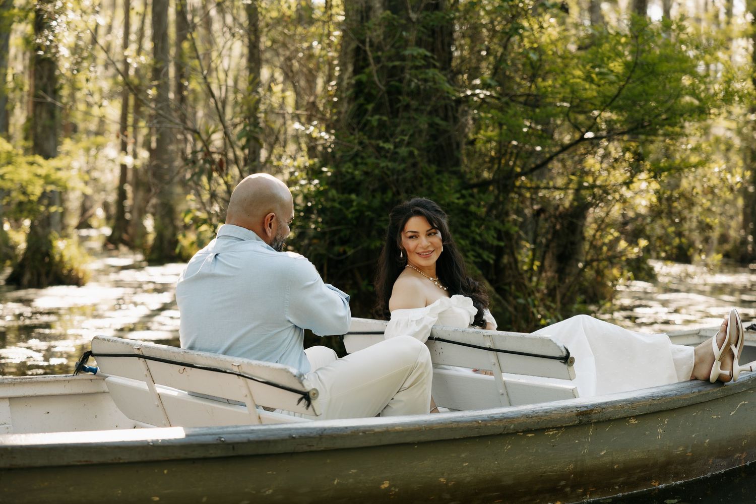 Couple in wedding attire sharing a romantic boat ride through a lush, sunlit cypress swamp forest.