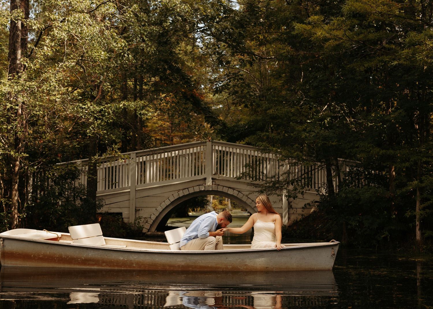 A couple in wedding attire shares a romantic boat ride on a serene lake with a white arched bridge surrounded by lush trees.