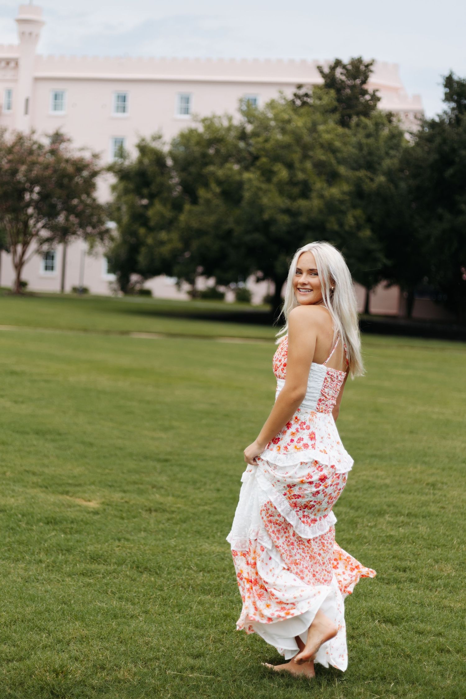 Woman in a flowing white and red floral dress smiles outdoors on a lawn with a pink building in the background.