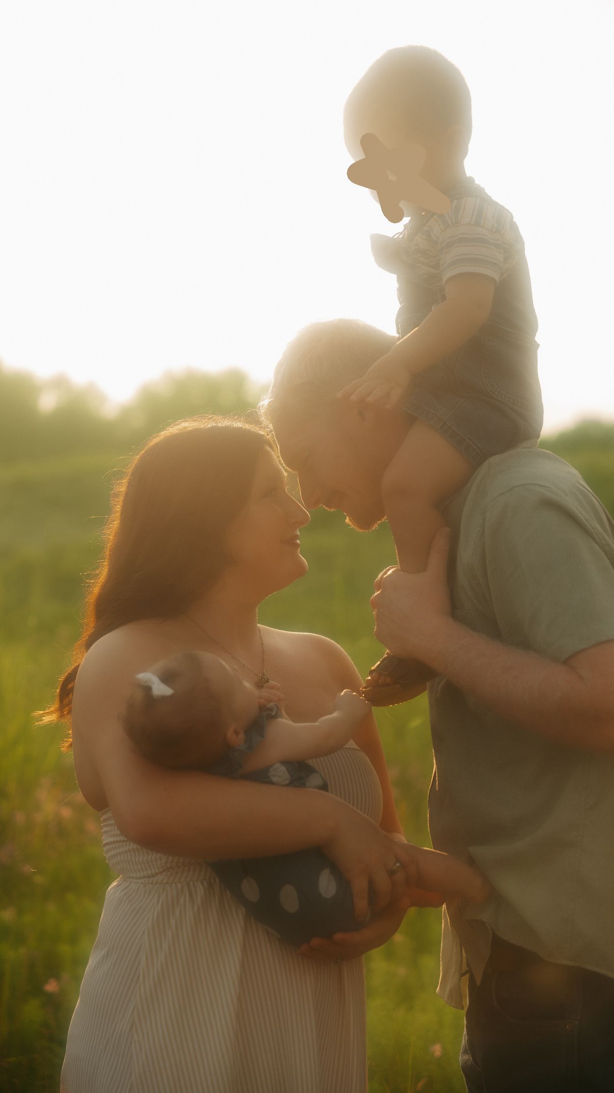 A young family shares a tender moment at golden hour in a field, parents touching foreheads while holding their two babies.