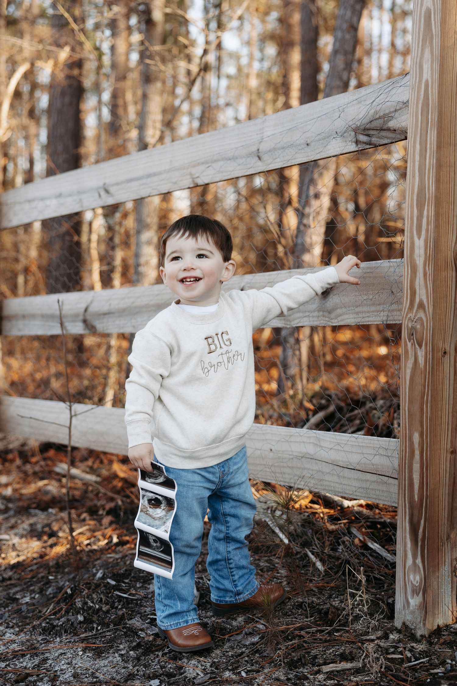 A child in jeans and white sweater plays and smiles along a wooden fence in an autumn forest setting.