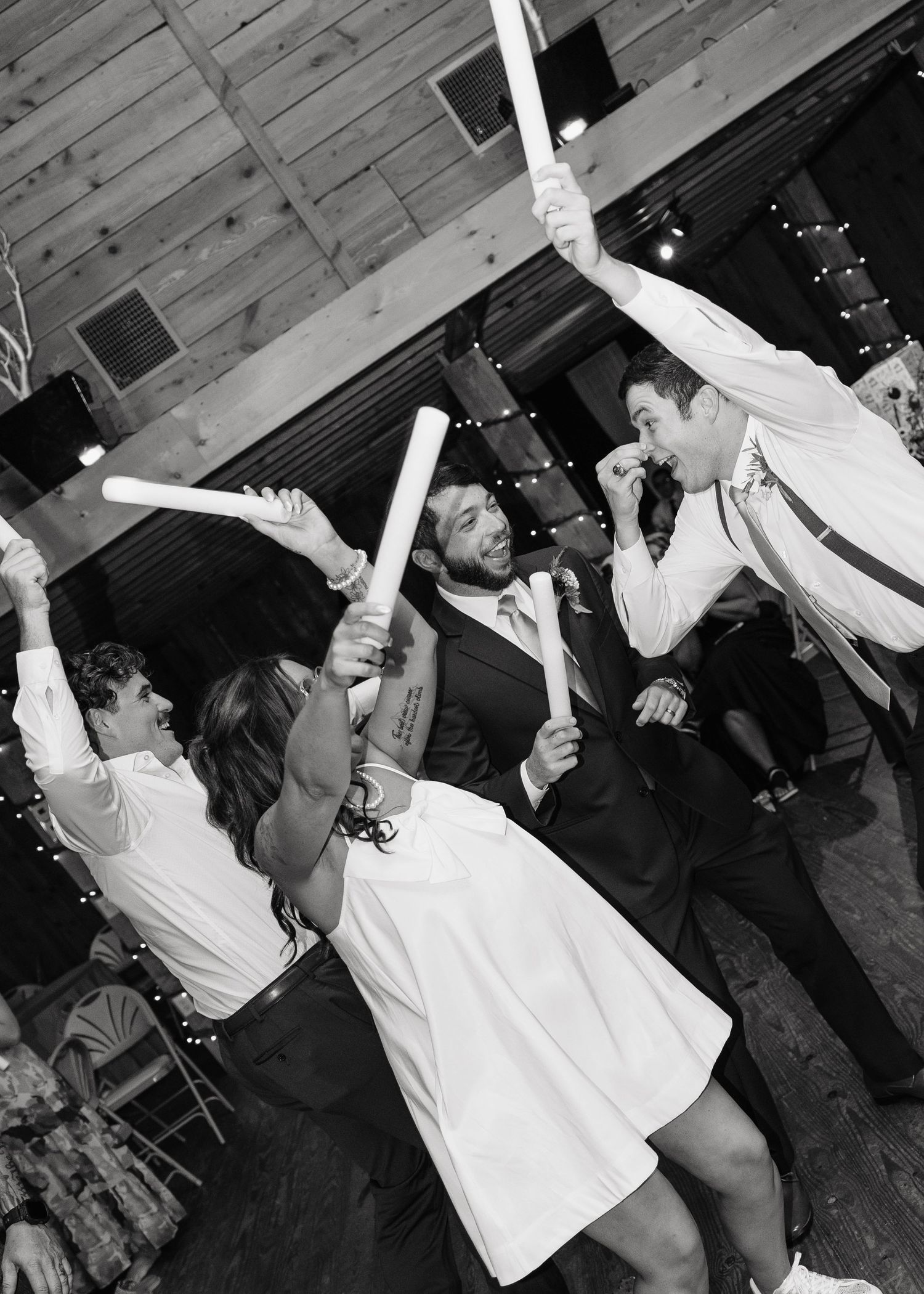 Black and white photo of wedding guests dancing and celebrating with glow sticks at a rustic barn reception.