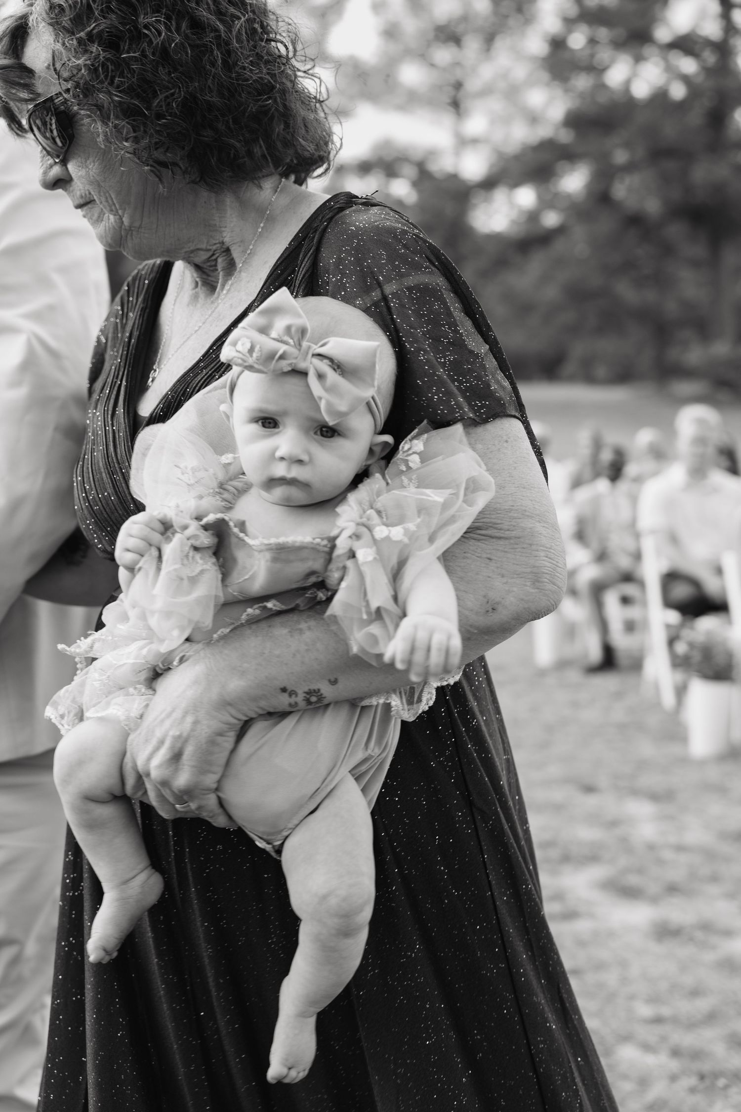 Black and white photo of a woman holding a baby girl wearing a bow headband at an outdoor wedding ceremony.